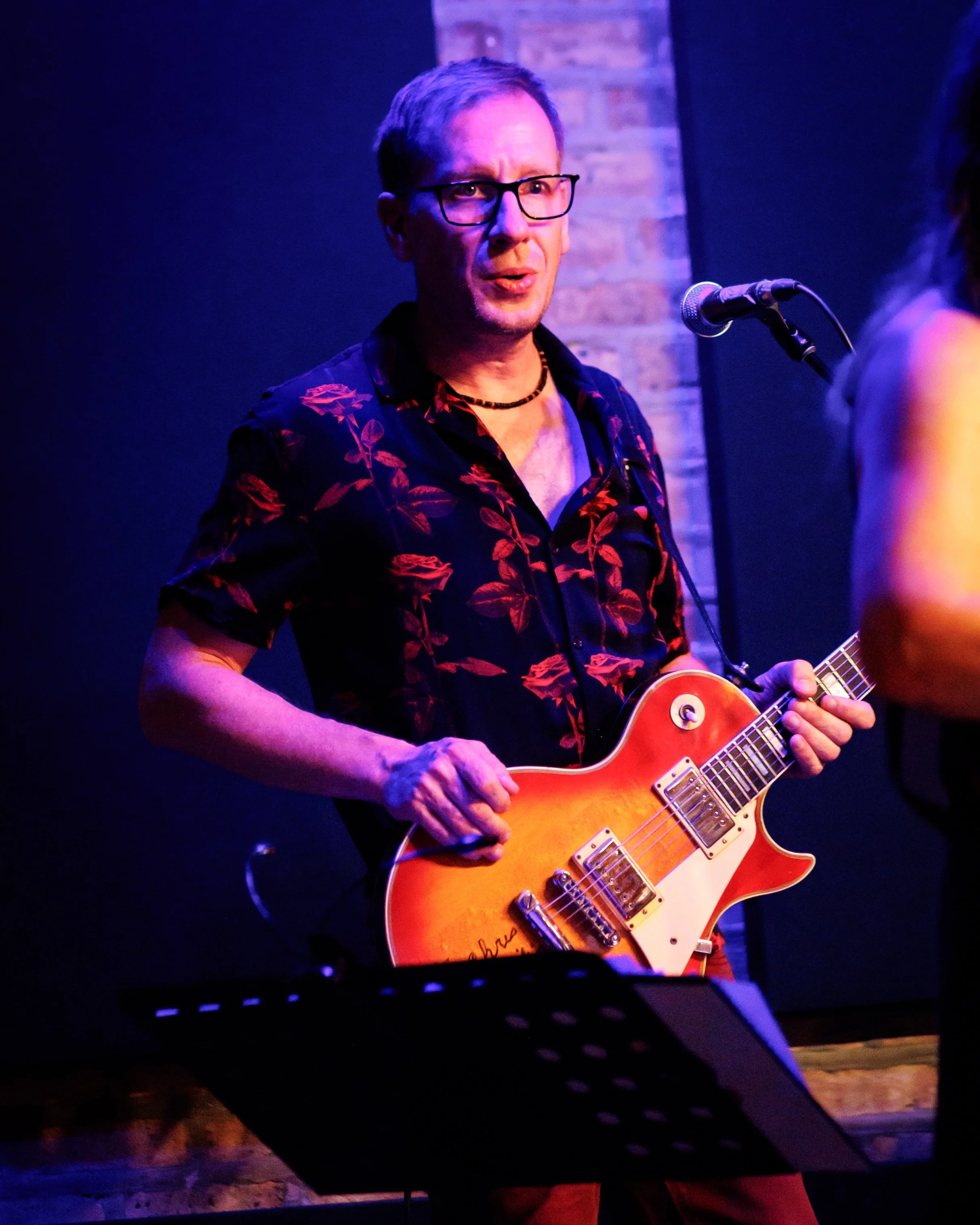 A man with glasses and a black floral shirt playing an orange electric guitar on stage with a microphone in front of him.