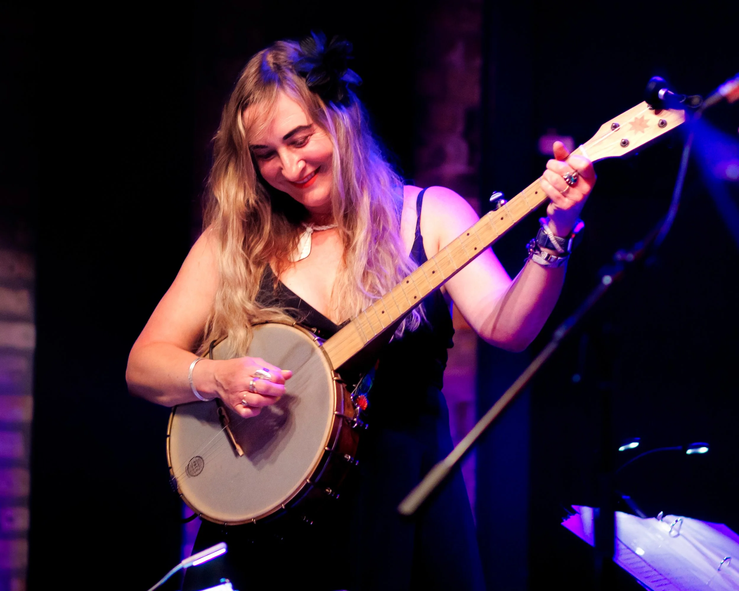 Woman playing banjo on stage, smiling, with piano nearby