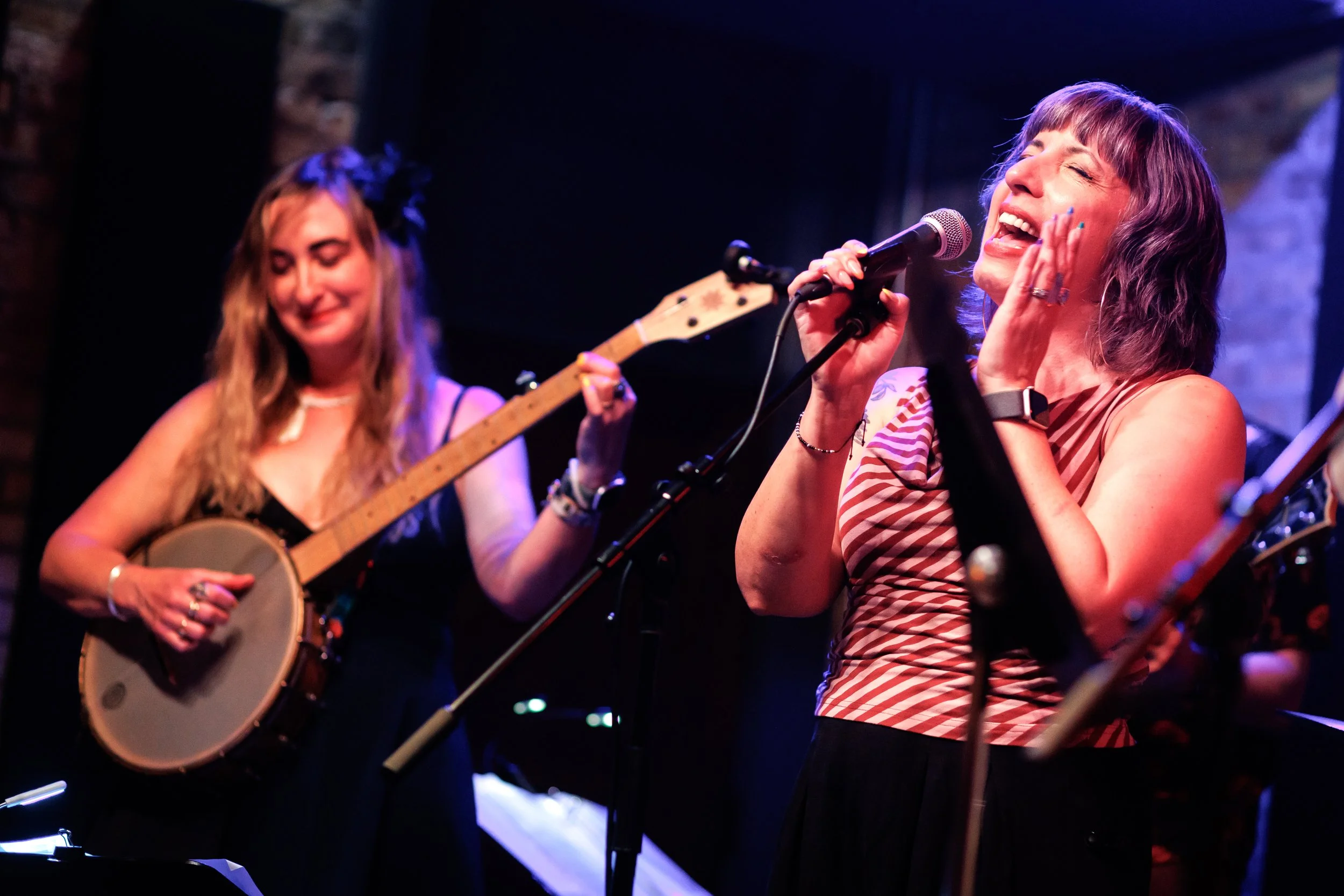 Two women performing music on stage; one singing into a microphone, smiling and touching her face, the other playing a banjo and smiling.