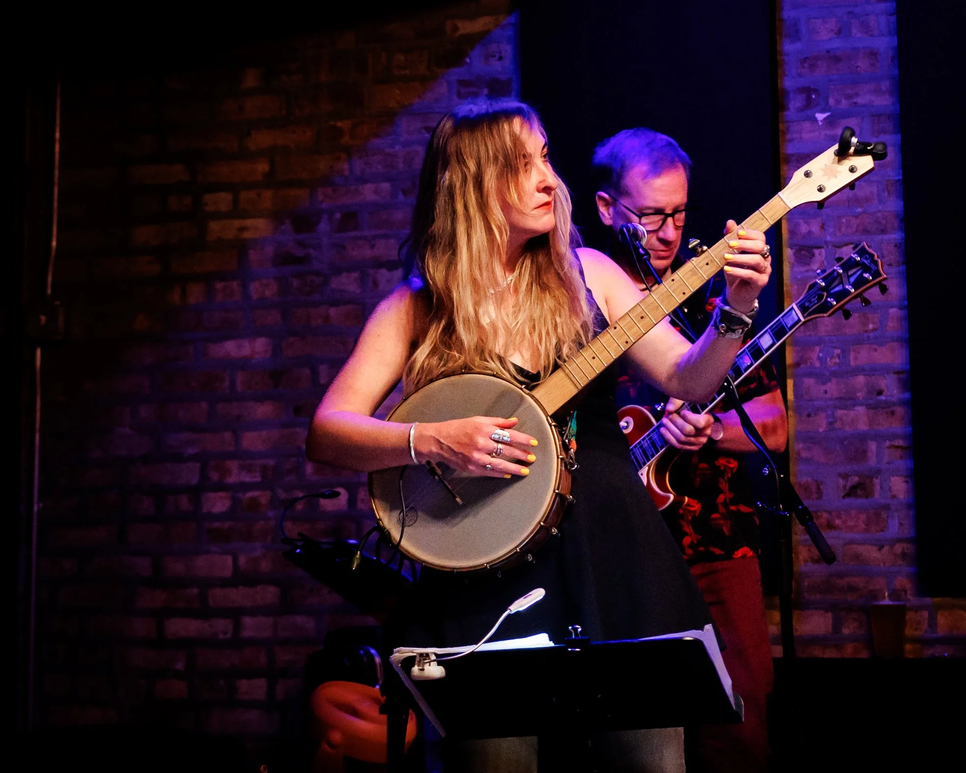 Two musicians playing acoustic and electric guitars on stage with brick wall background.