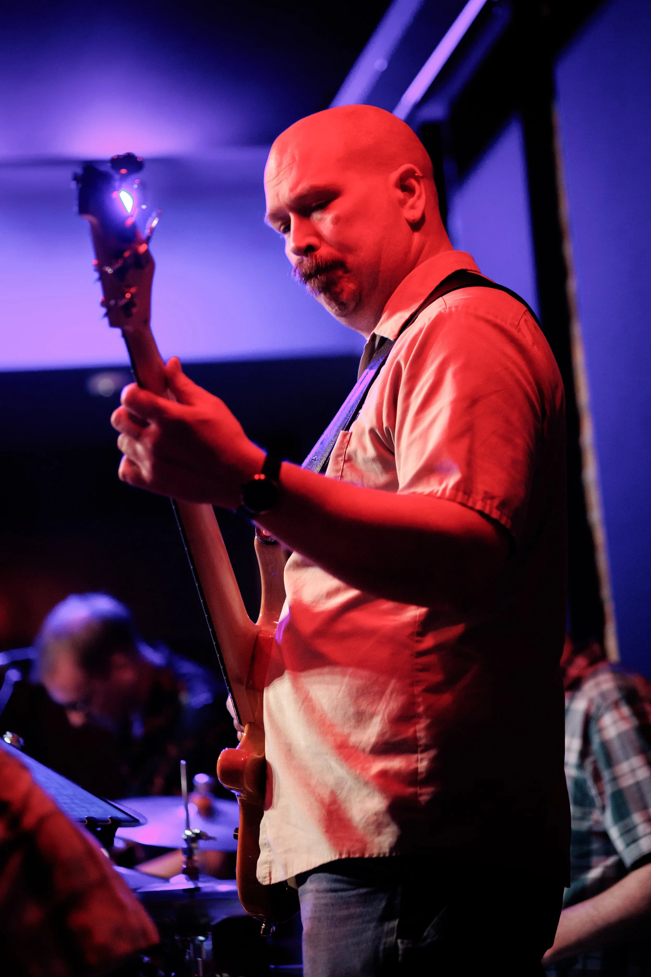 Man with a bald head and mustache playing an electric guitar in a dimly lit room with purple and blue stage lights, with other band members visible in the background.