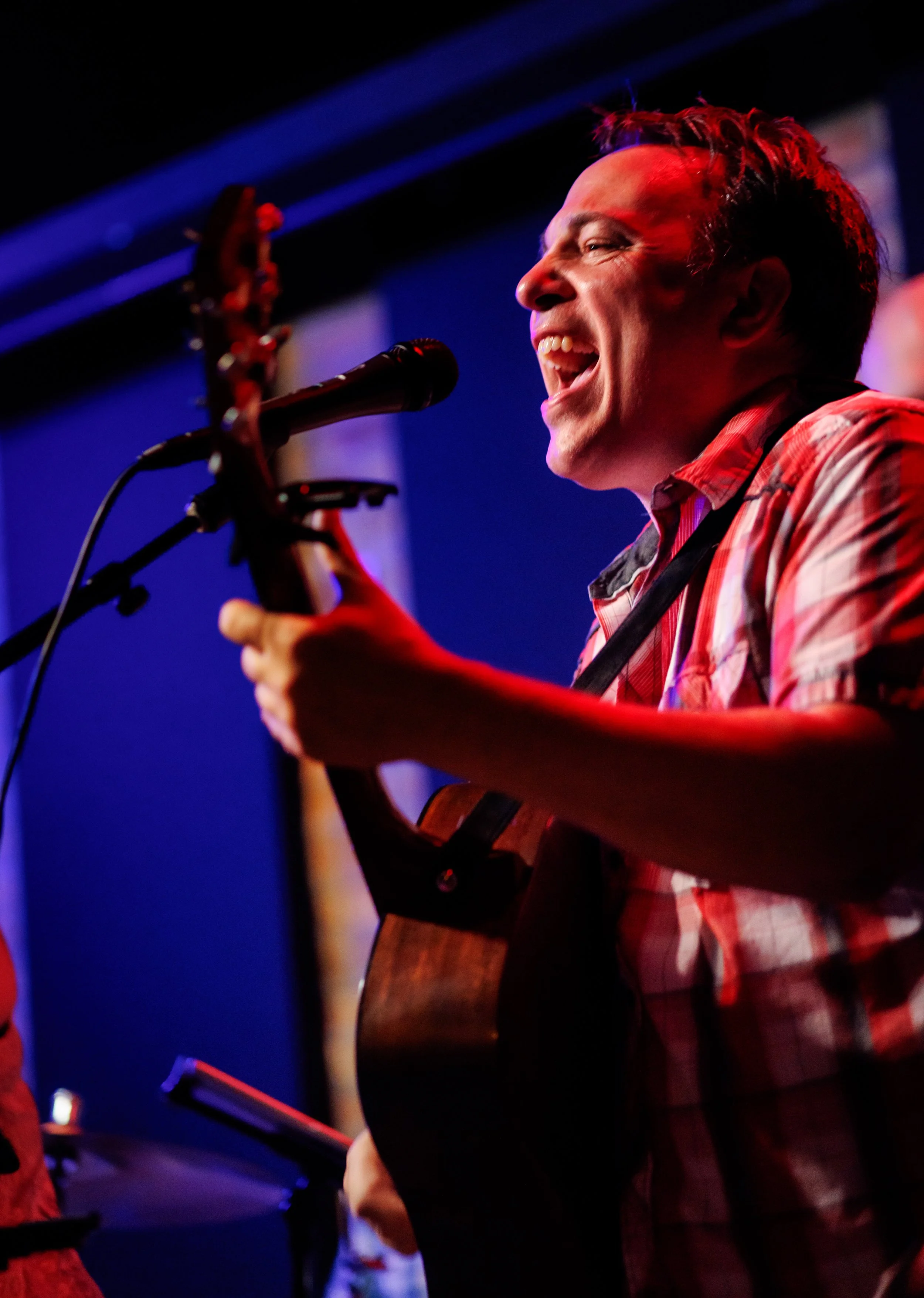 A man singing passionately while playing an acoustic guitar on stage, with a microphone in front of him, illuminated by stage lighting.
