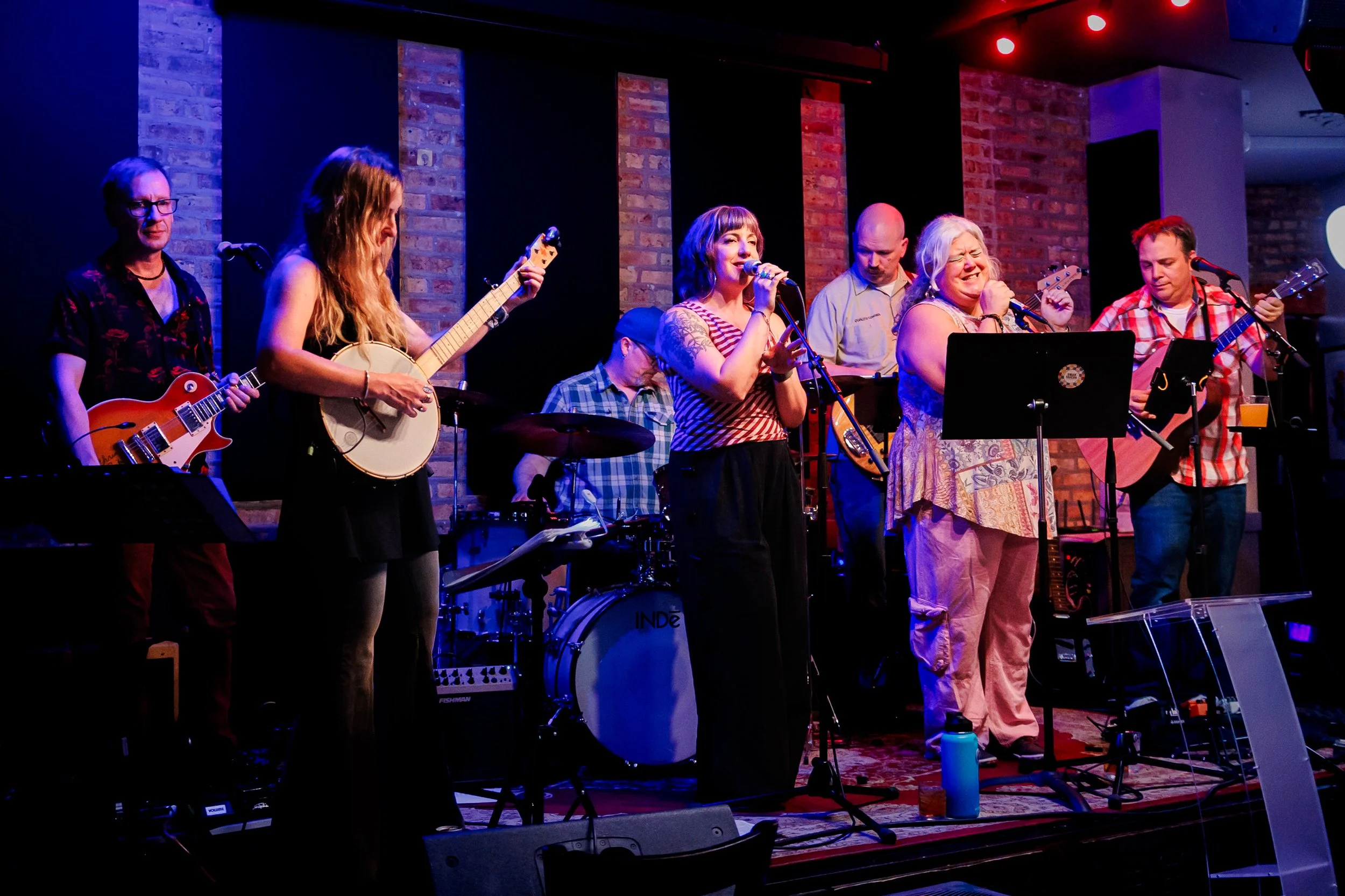 A group of seven musicians performing on a stage with brick wall background, including two women singing into microphones, one woman playing a banjo, two men playing guitars, one man on drums, and one man on keyboard, lit with stage lights.