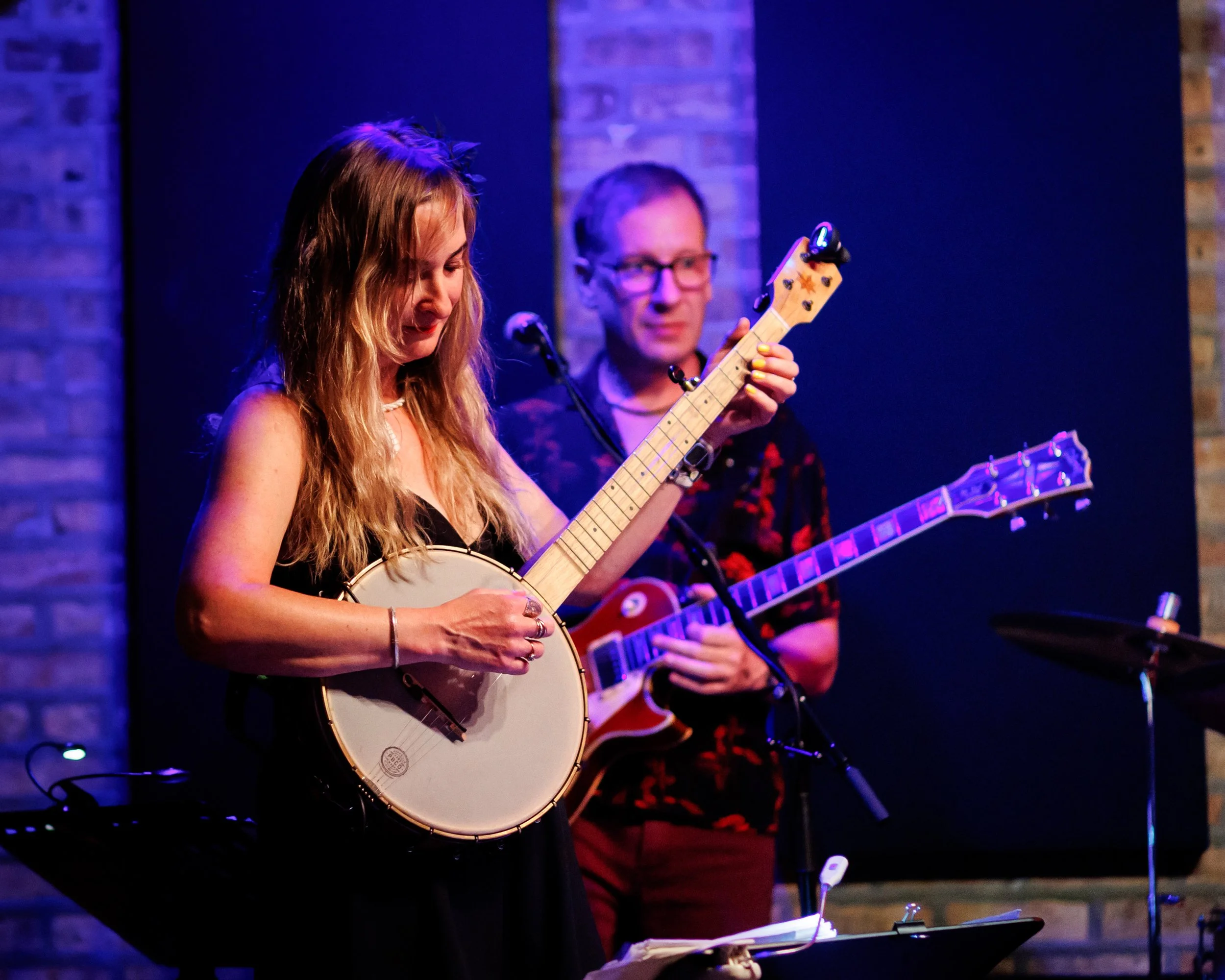 A woman playing a banjo on stage with a man playing a guitar in the background.