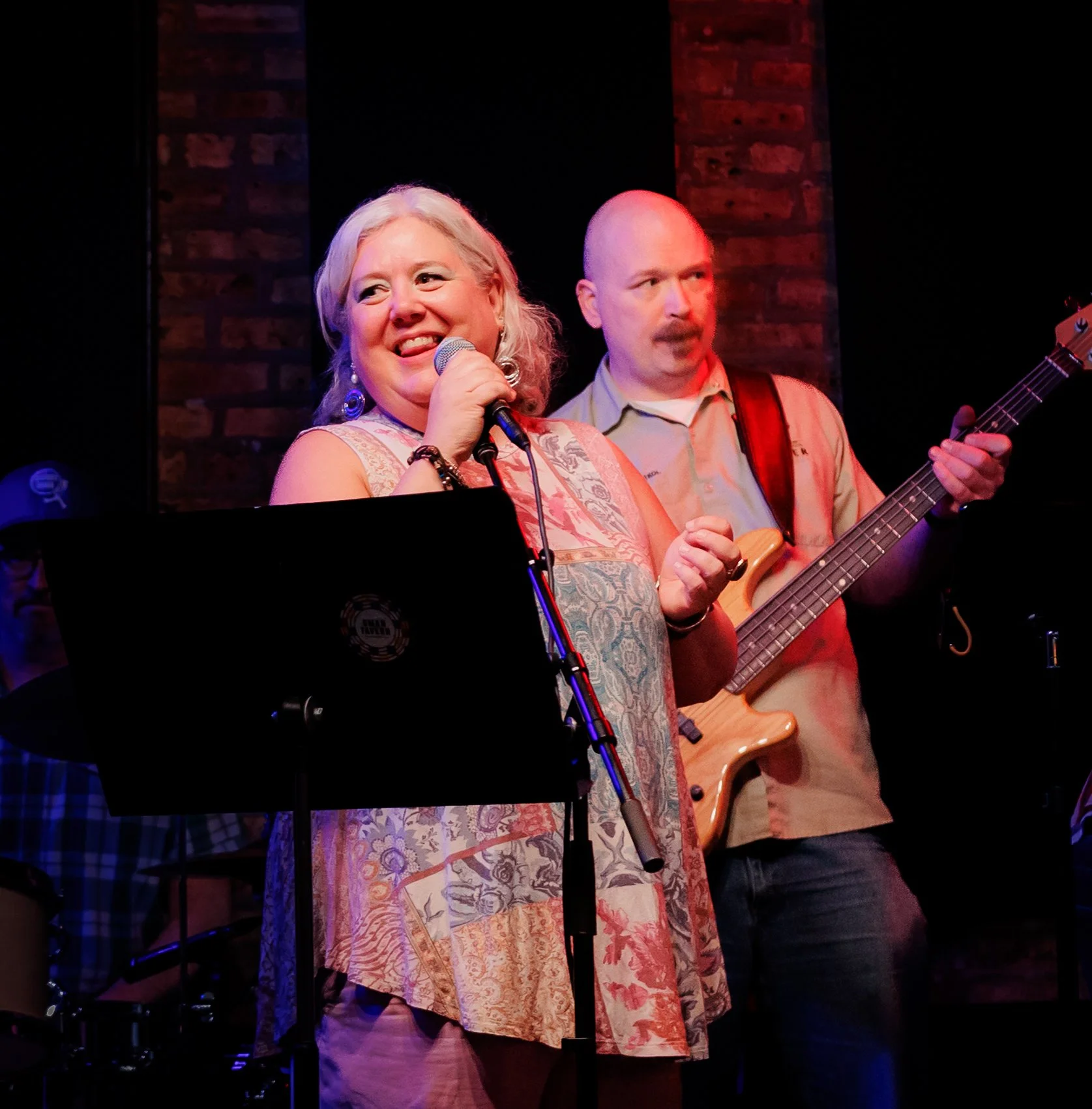 A group of musicians performing on stage at a venue with brick walls. A smiling woman with gray hair, wearing a colorful sleeveless dress, is singing into a microphone. A man next to her, with a bald head, is playing an electric bass guitar.