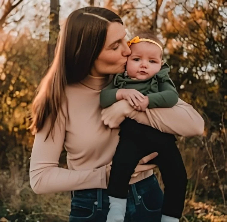 A mother with long brown hair holding her daughter outdoors during autumn, with trees and fall foliage in the background.
