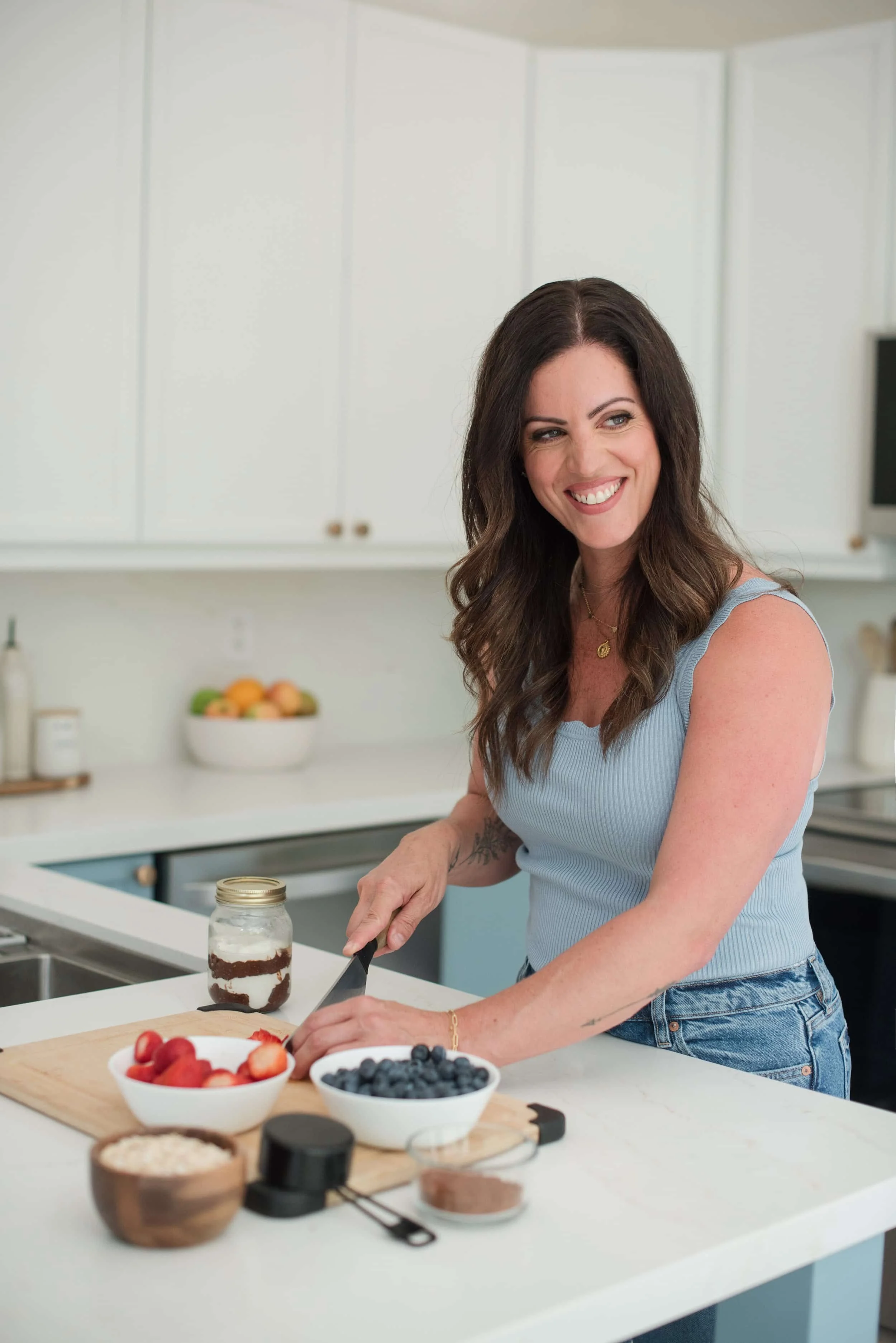 A woman with long brown hair smiles while preparing food in a modern kitchen. She is wearing a light blue sleeveless top and jeans, with bowls of strawberries, blueberries, and other ingredients on the white countertop.