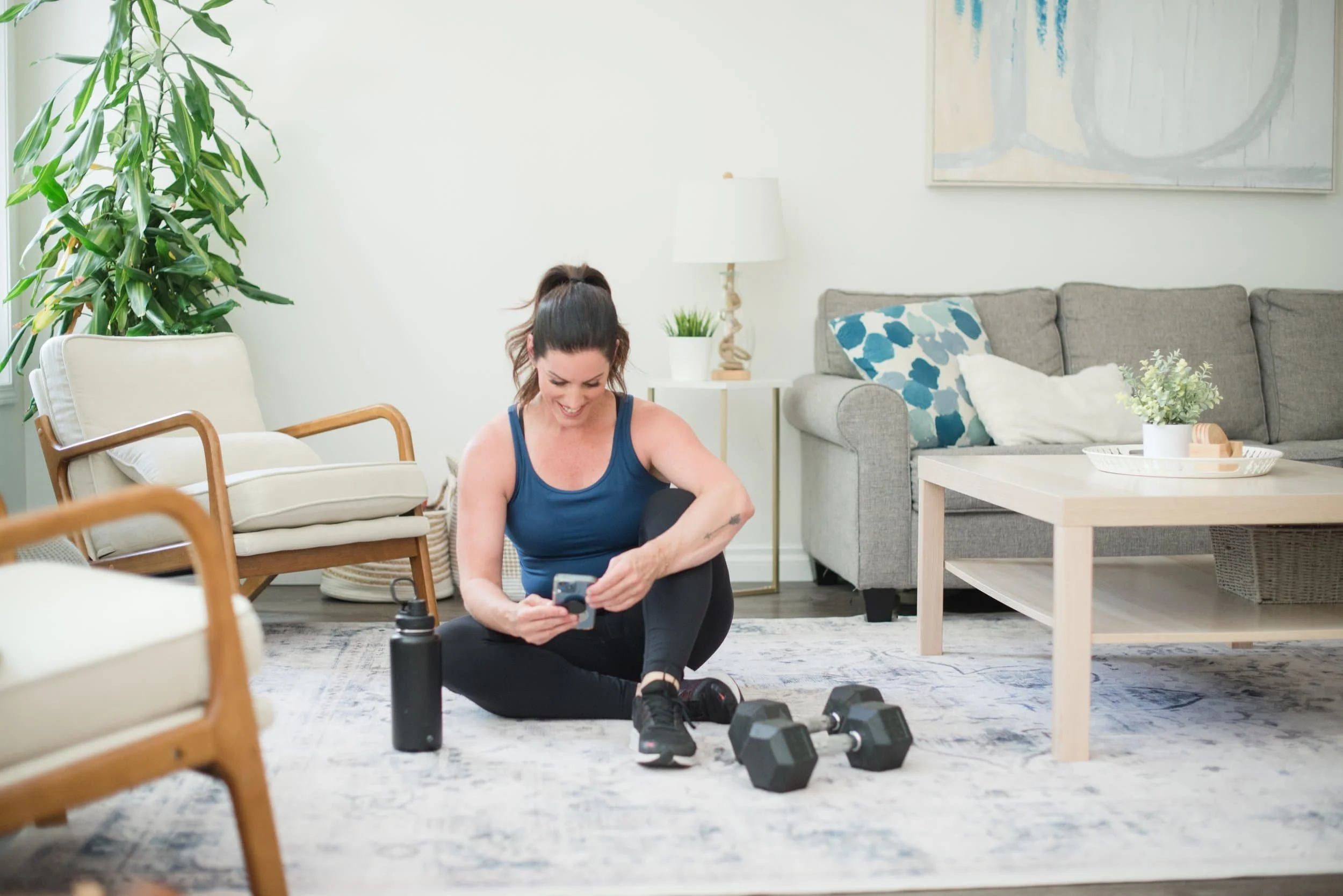 A woman sitting on the floor in a bright living room, smiling while looking at her phone, with gym equipment including dumbbells and water bottles nearby.