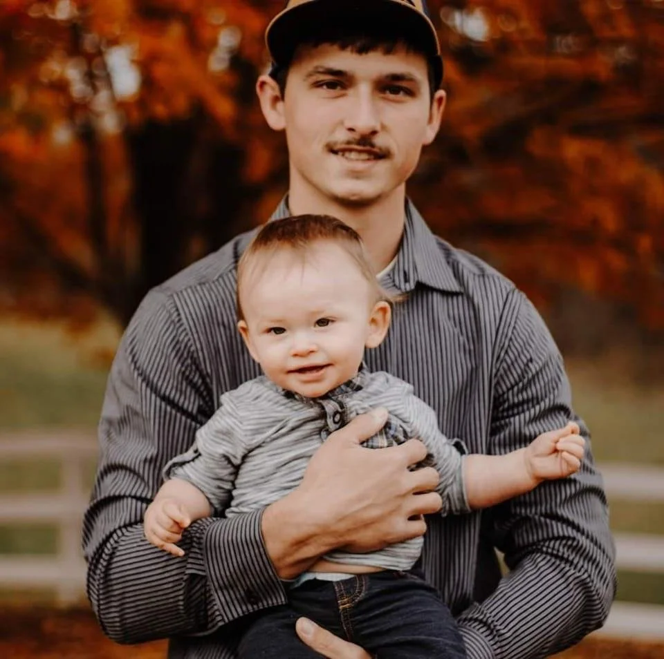 A man with a mustache and wearing a striped gray shirt holds a young toddler with light hair and a striped shirt outdoors with autumn trees in the background.