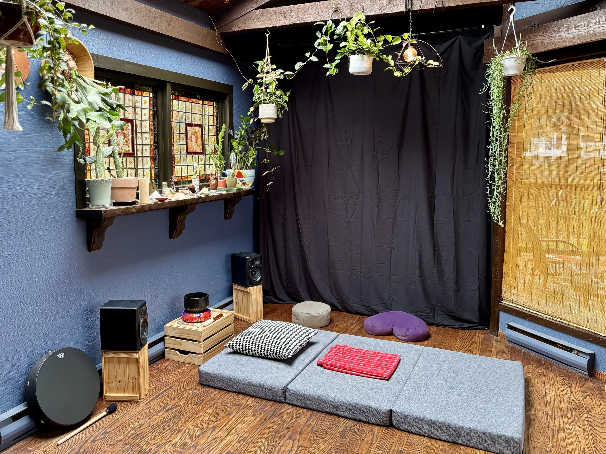 A cozy meditation or yoga space with blue walls, wooden flooring, and black curtains. There are plants hanging from above, a small window with stained glass, and a shelf with decorative items. On the floor, there are gray foam mats, cushions, and a folded red blanket.