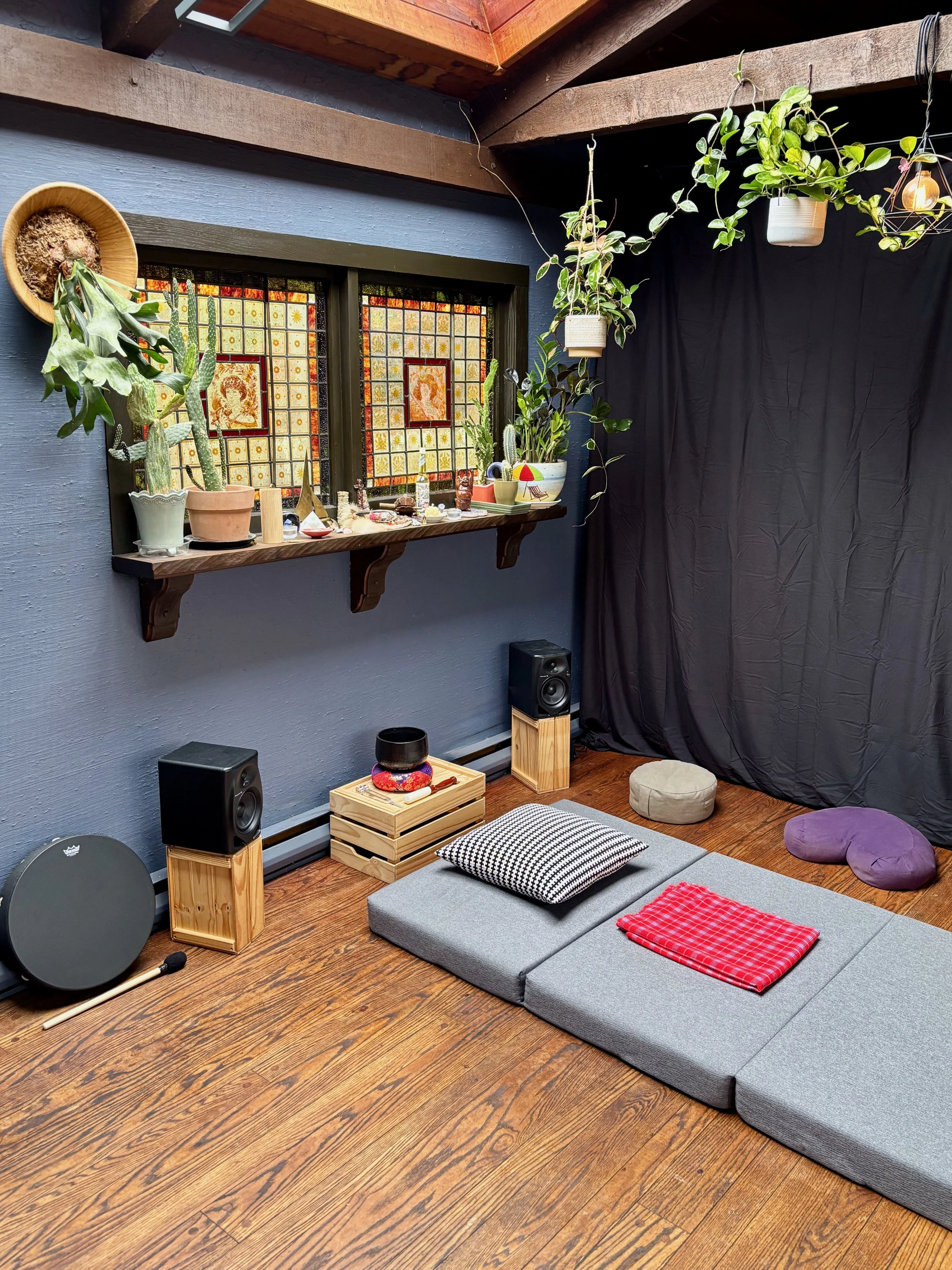A cozy meditation or yoga space with a wooden floor, gray foam mats, cushions, and blankets. The space features a wall with decorative stained glass windows, plants, and hanging lights. There's a shelf with plants and speakers. Victoria BC 5-MeO-DMT.