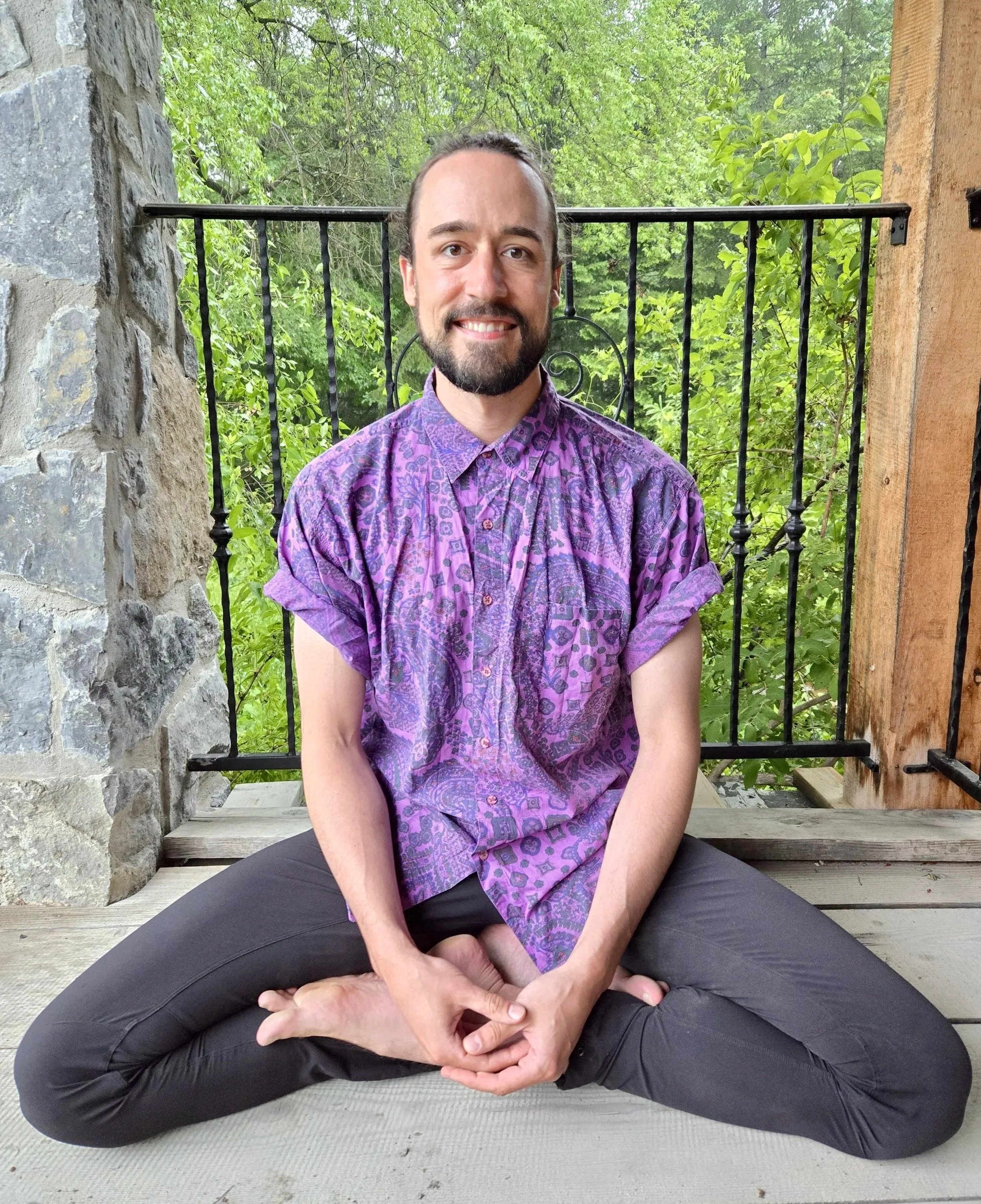 A man with a beard sitting cross-legged on a wooden porch, smiling at the camera, wearing a purple patterned short-sleeve shirt, with a background of green trees and foliage. Brendon Campbell. Cosmic Otter. Psychedelic Facilitator. 5-MeO-DMT Guide.