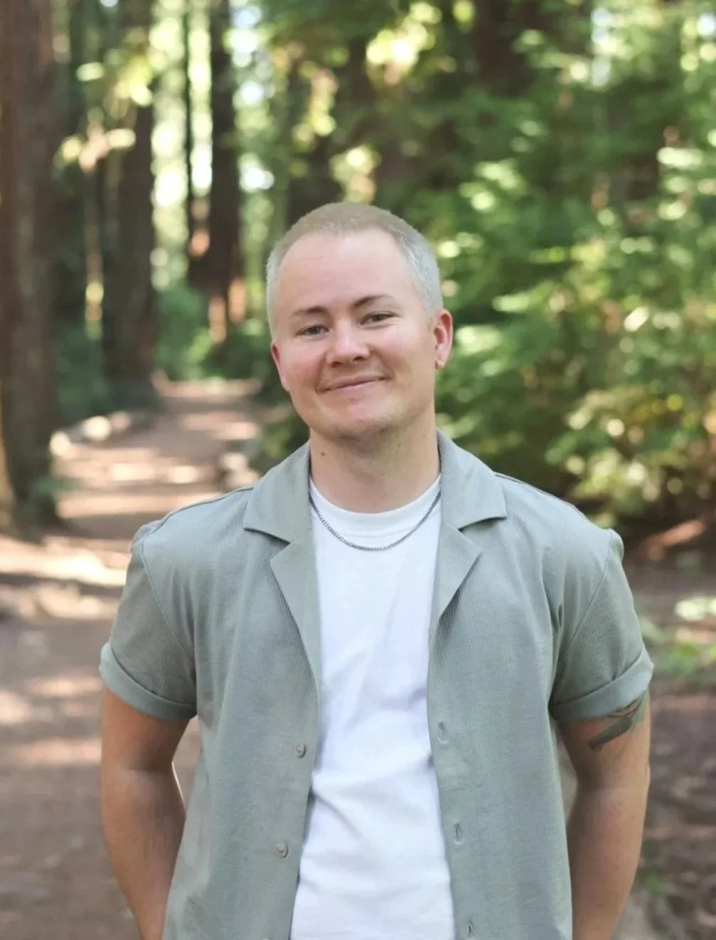 A white, 30-something transmasculine nonbinary person stands calmy on a trail in the redwoods. The photo shows their torso, and a blurred background of a nature path surrounded by redwoods.