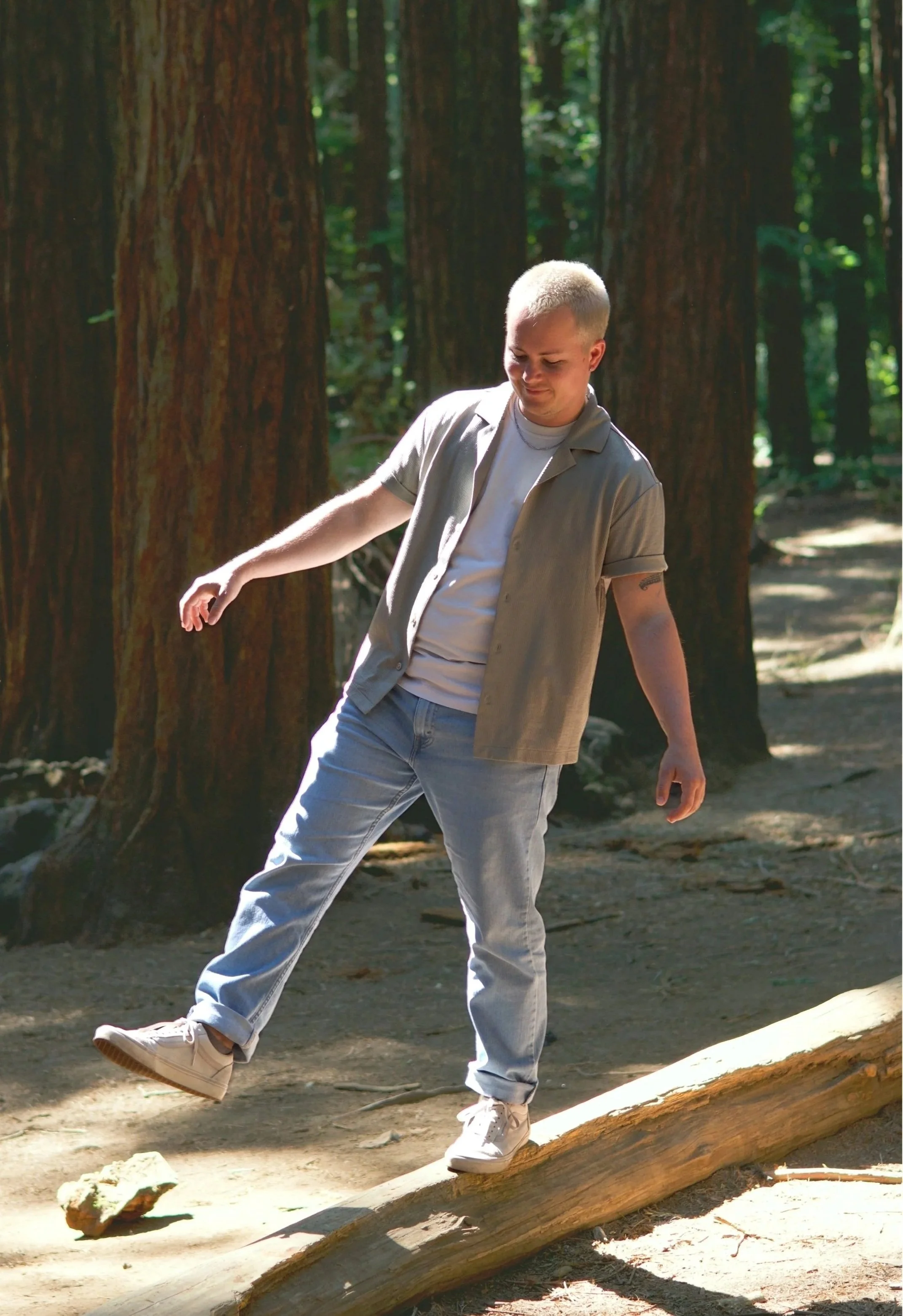 A white, 30-something transmasculine nonbinary person with short bleached hair is walking on a fallen tree in the redwoods. He is wearing a white t-shirt, an unbuttoned sage green short sleeve button up, light blue jeans, and casual white shoes.