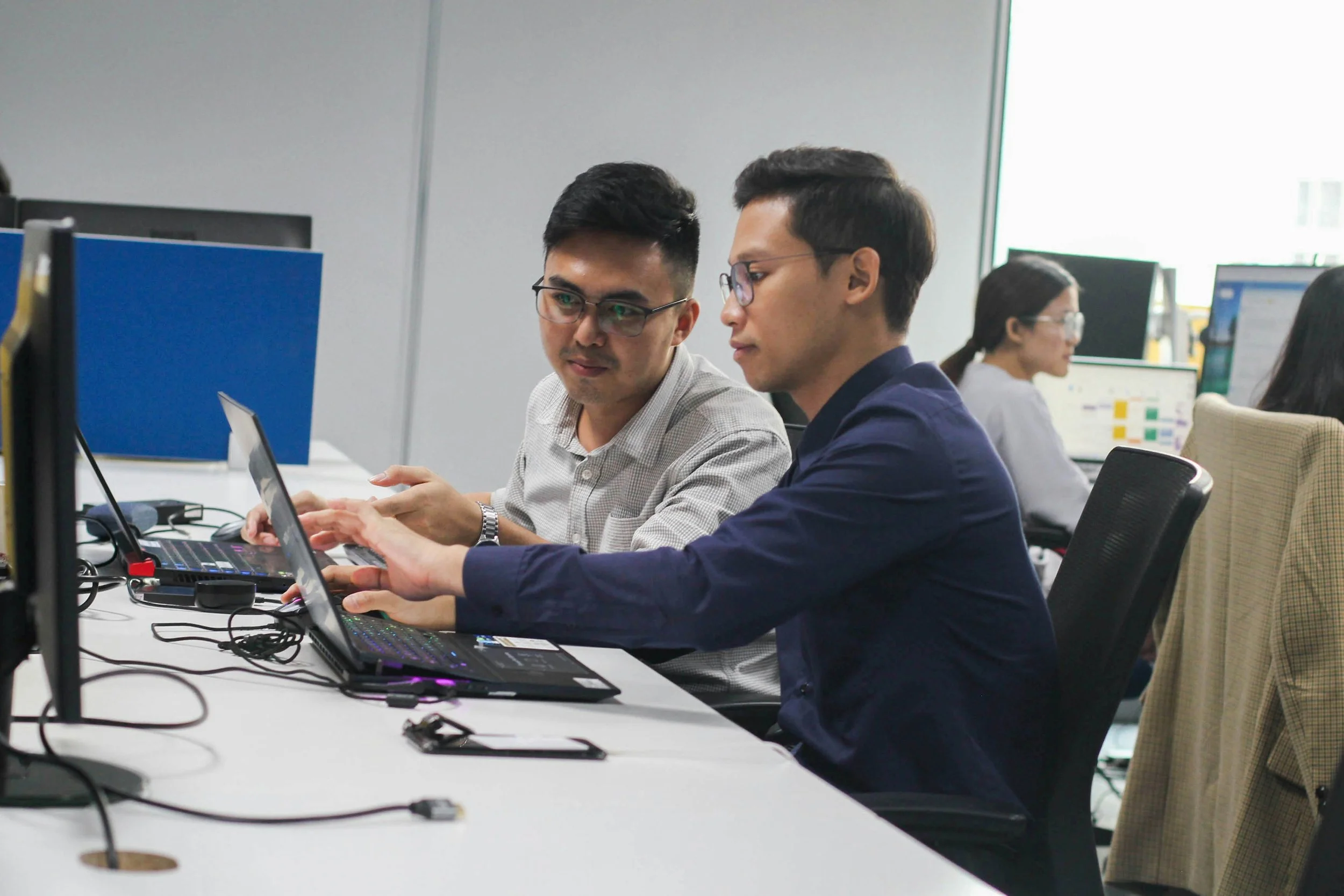 Two men in an office working with laptops, one showing the other something on the screen, with two women working in the background.