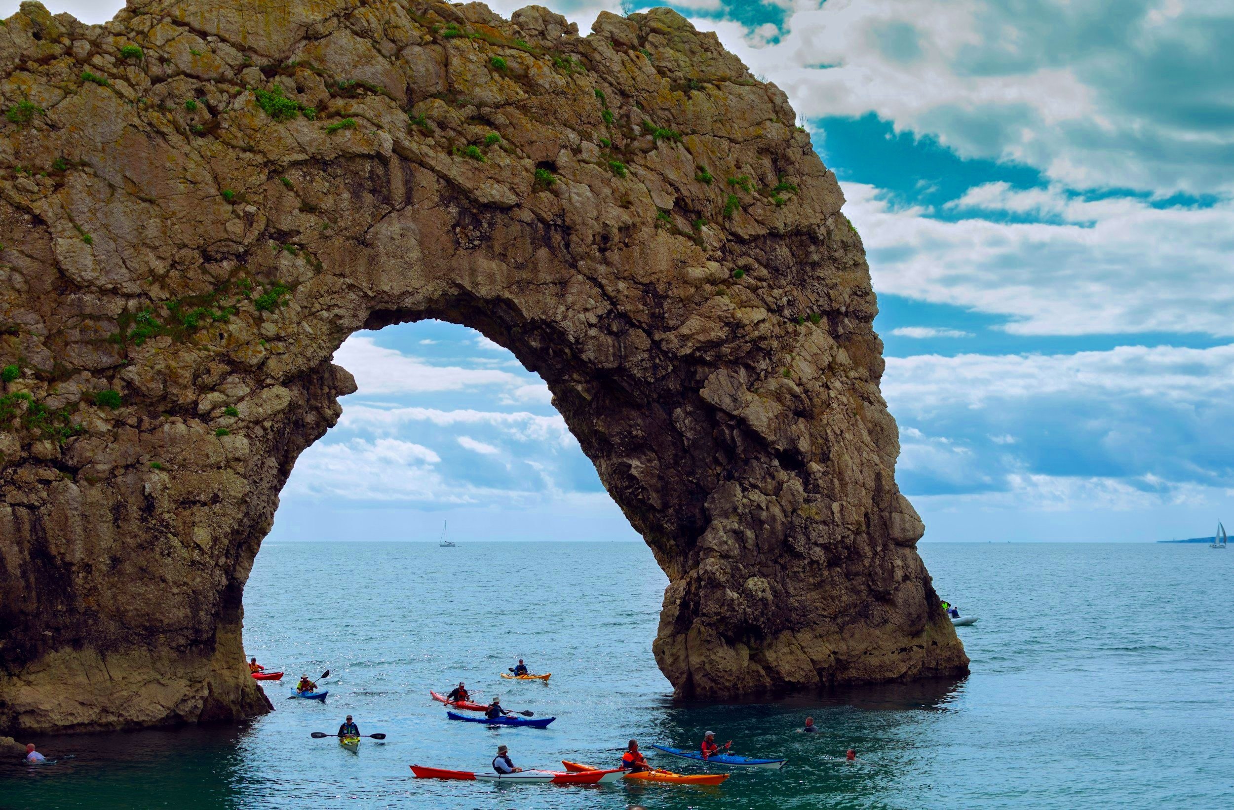 People kayaking underneath a large natural rock arch formation in the ocean with a partly cloudy sky.