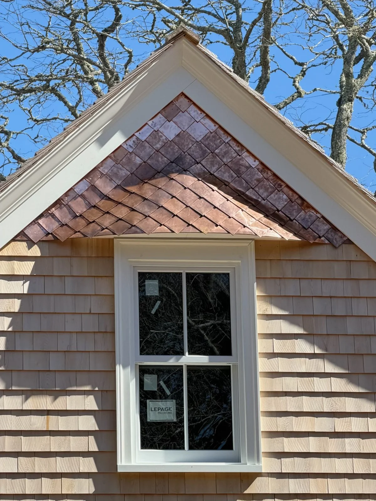 Close-up of a house exterior with beige siding, a window with white trim, and a gable roof with brown shingles. The sky is clear and blue, with tree branches in the background.