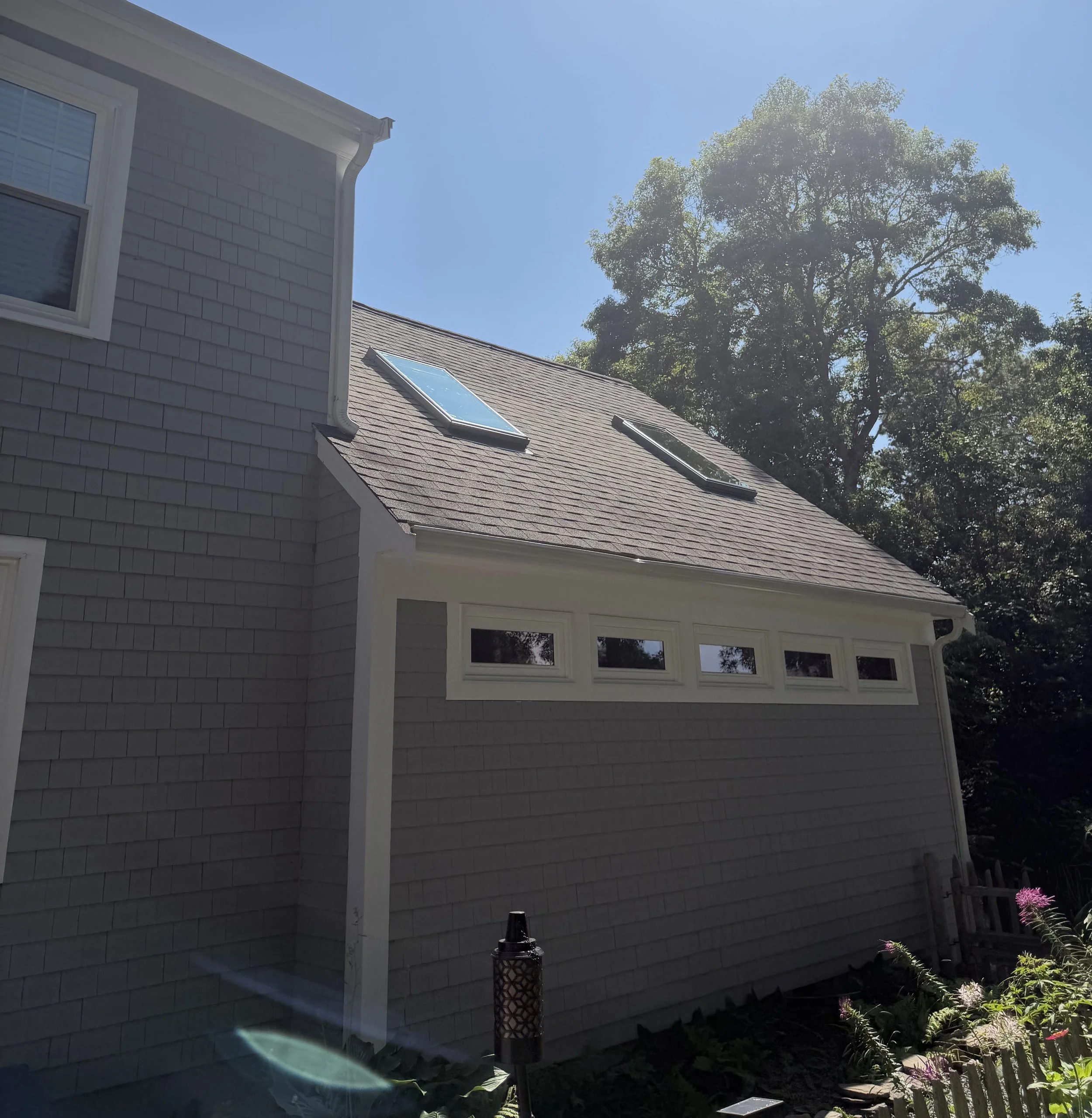 A house with gray siding and a sloped roof with two skylights, set against a bright blue sky and a large leafy tree. There is a black and gold decorative lantern in the garden area underneath the house's window, which has white trim.