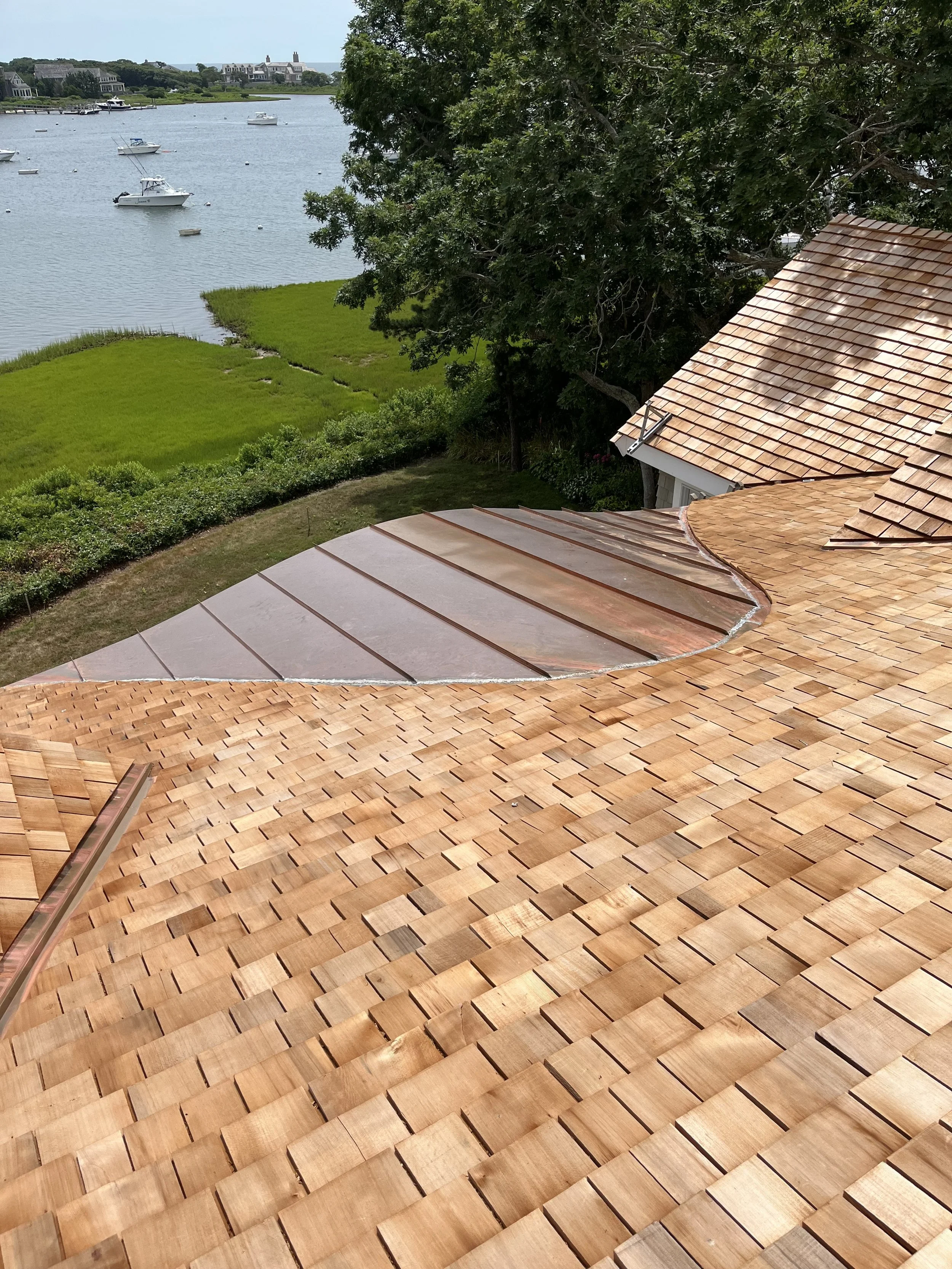 View from a rooftop with wooden shingles overlooking a grassy area, trees, and a river with boats in the background.