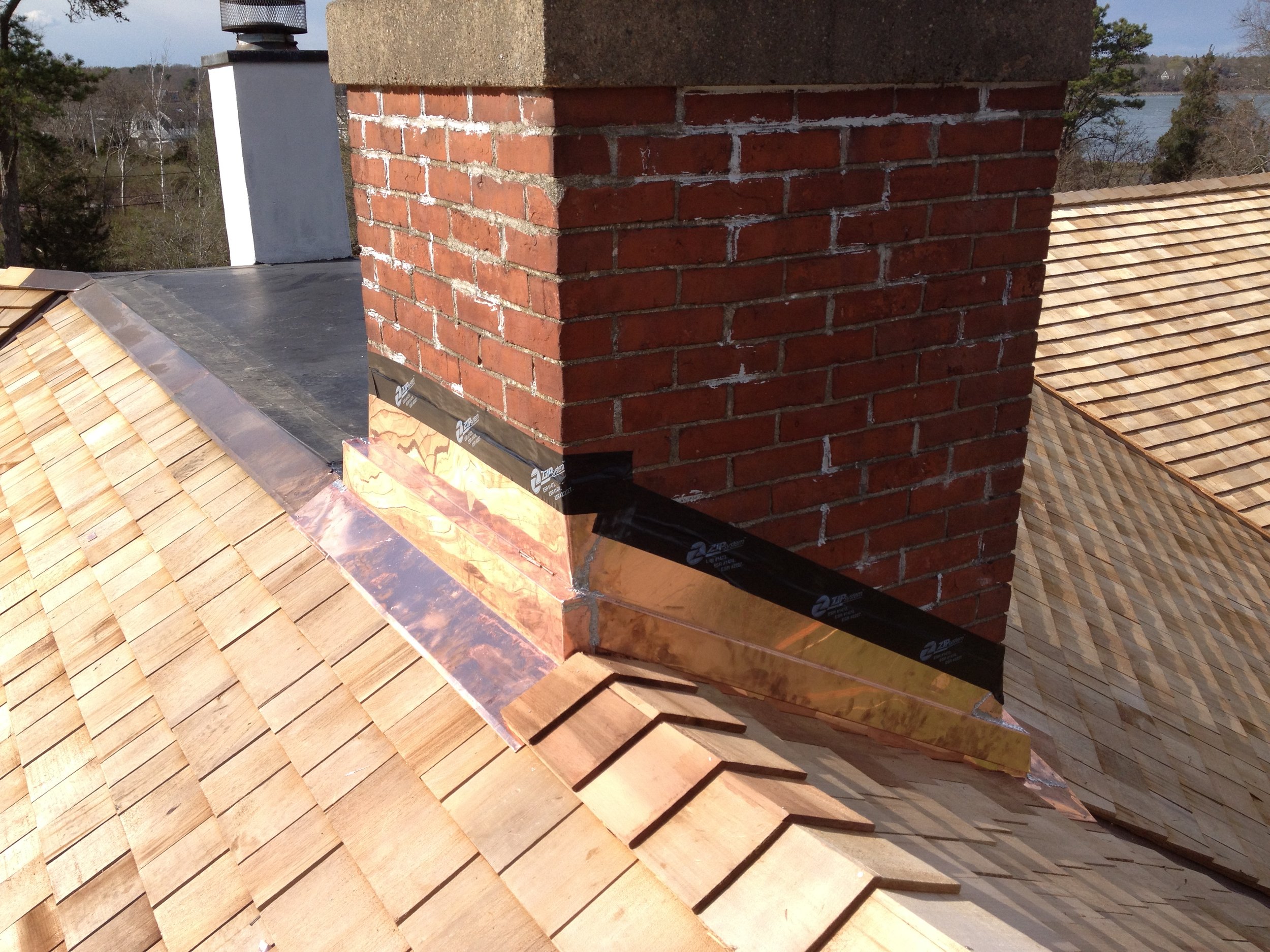A rooftop with a brick chimney, new copper flashing, and different roofing materials including shingles and metal flashing. The background shows trees and a body of water.