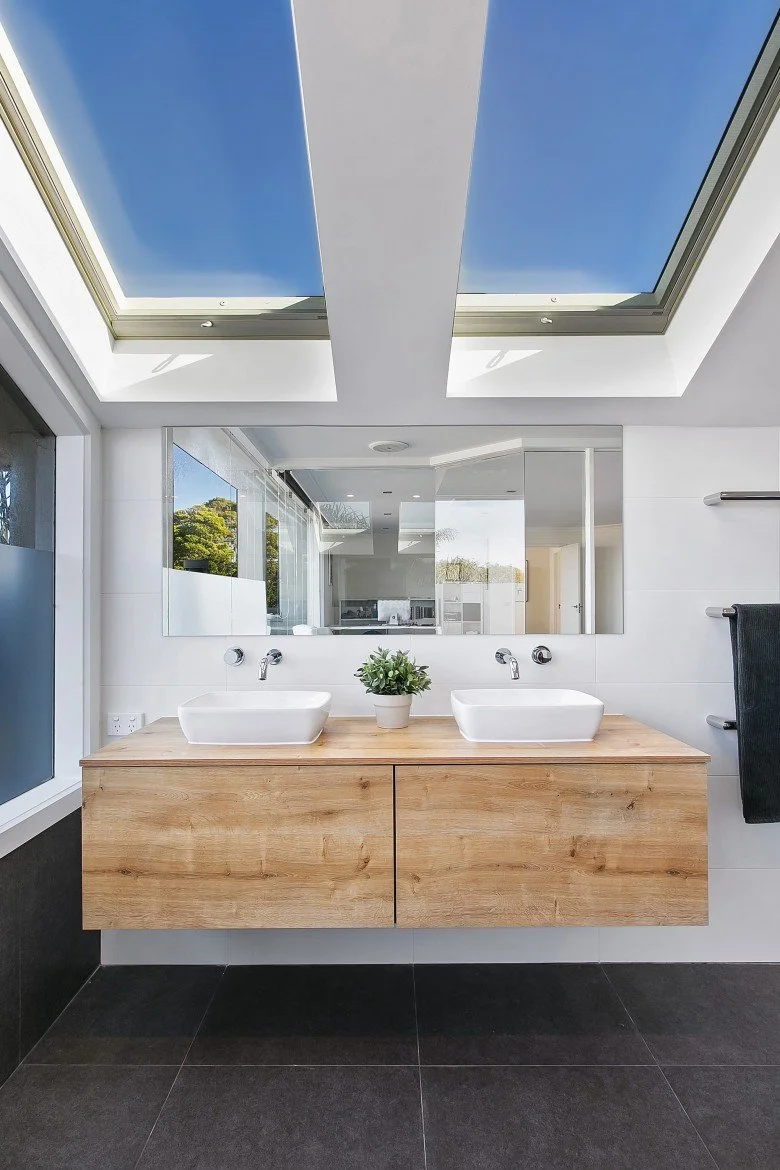 Modern bathroom with a wooden vanity, two white vessel sinks, a large mirror, a skylight showing blue sky, and a potted plant on the vanity.