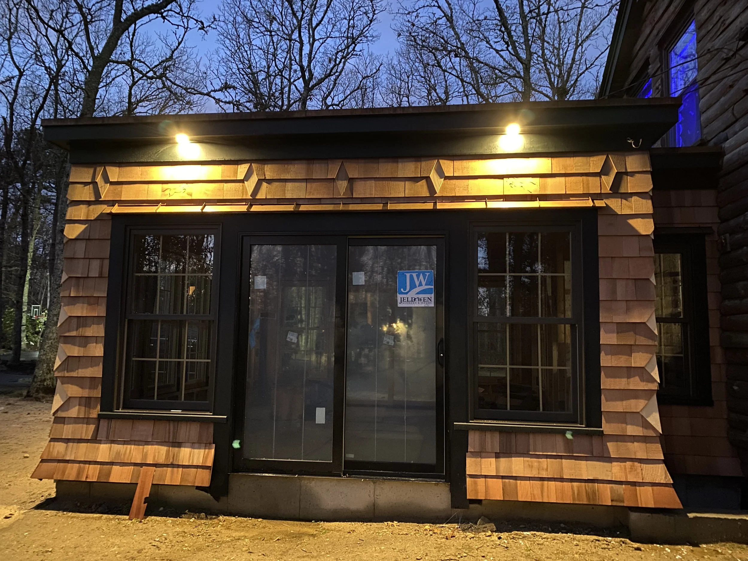 Front view of a building with a shingled exterior, glass sliding door, and windows, illuminated by two overhead lights at dusk.