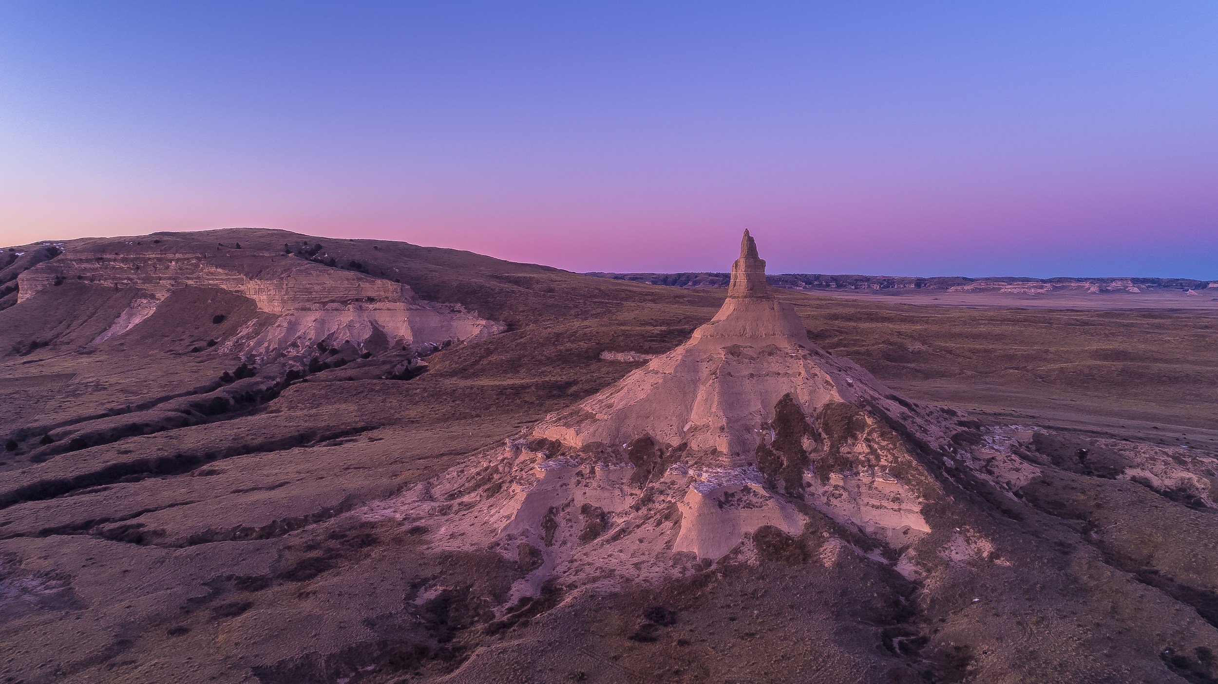 A landscape view of a purple and pink sunset over rocky, layered formations and a dome-shaped mountain with a pointed peak in the foreground.