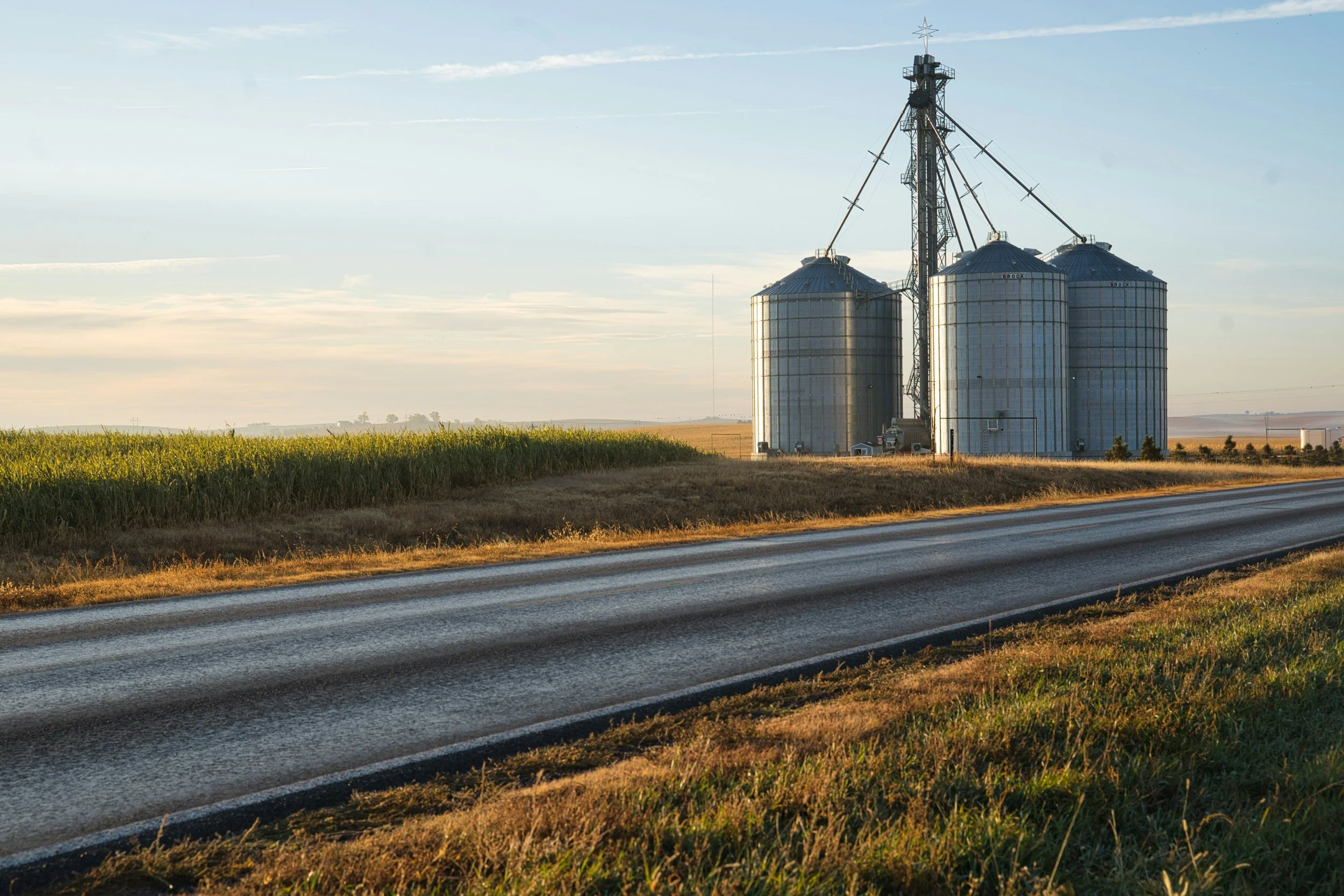 A rural landscape with a two-lane paved road in the foreground, green fields to the left, and a row of silos with a grain elevator in the distance under a clear sky.