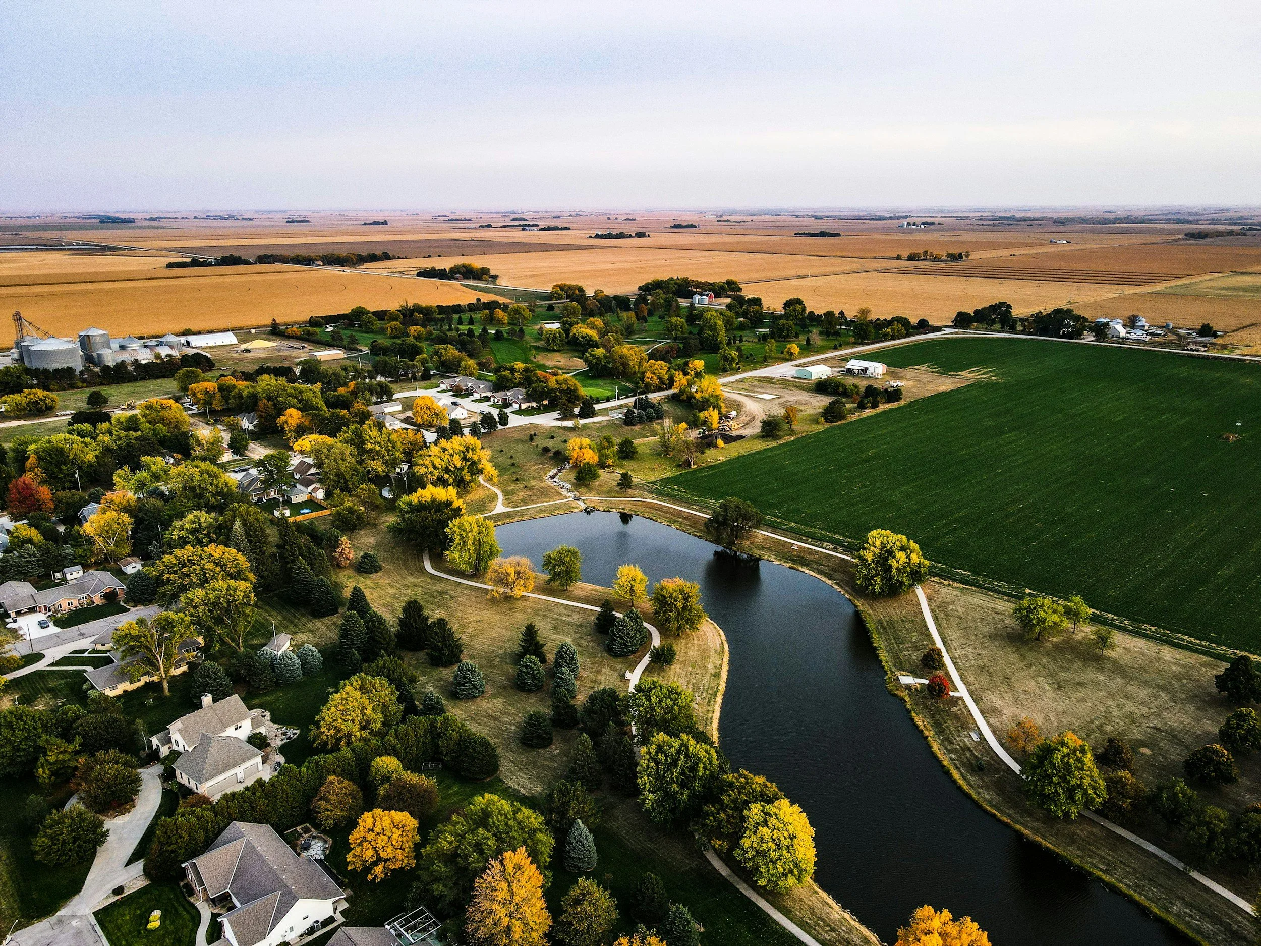 An aerial view of a rural landscape featuring a pond, residential houses, trees with fall foliage, and surrounding farmland, including large fields and farm buildings.
