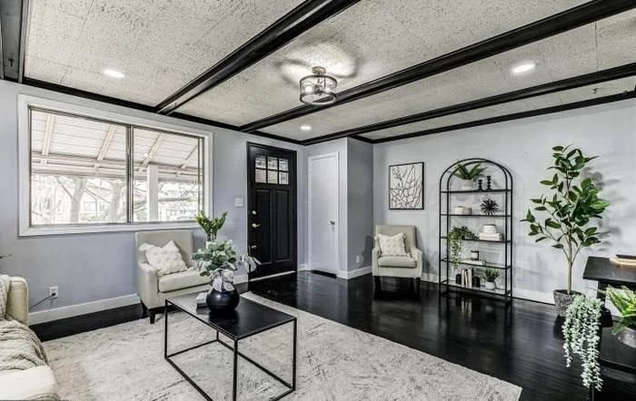Living room with black front door, large window, gray walls, black trim, light-colored armchairs, a black coffee table with a vase, dark hardwood floors, a white rug, and a black metal bookshelf with plants and decor.