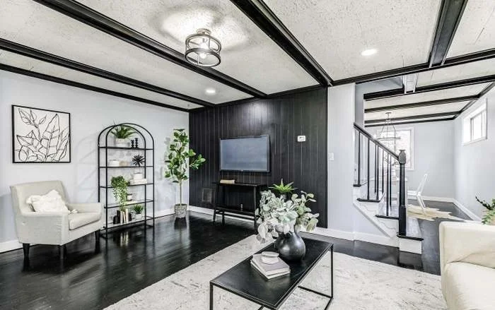 Modern living room with black and white decor, a wall-mounted TV, a beige armchair, black side table, bookshelf, potted plants, staircase, and ceiling lights.