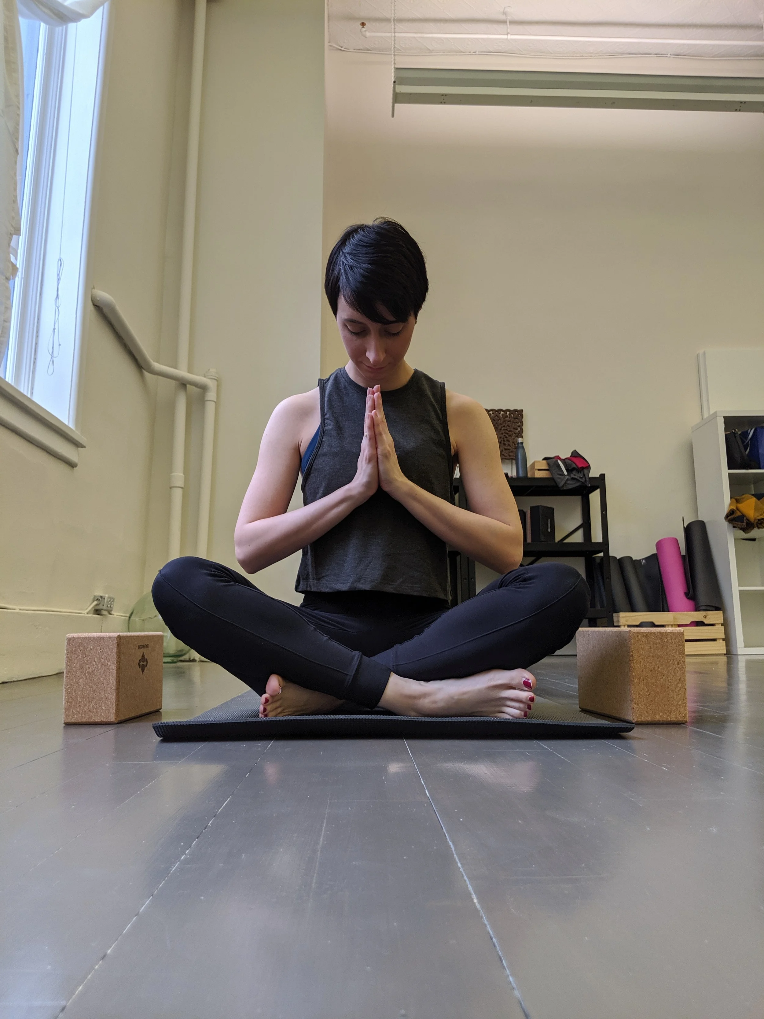 A woman practicing yoga indoors while sitting cross-legged on a mat with her hands in prayer position and eyes closed.