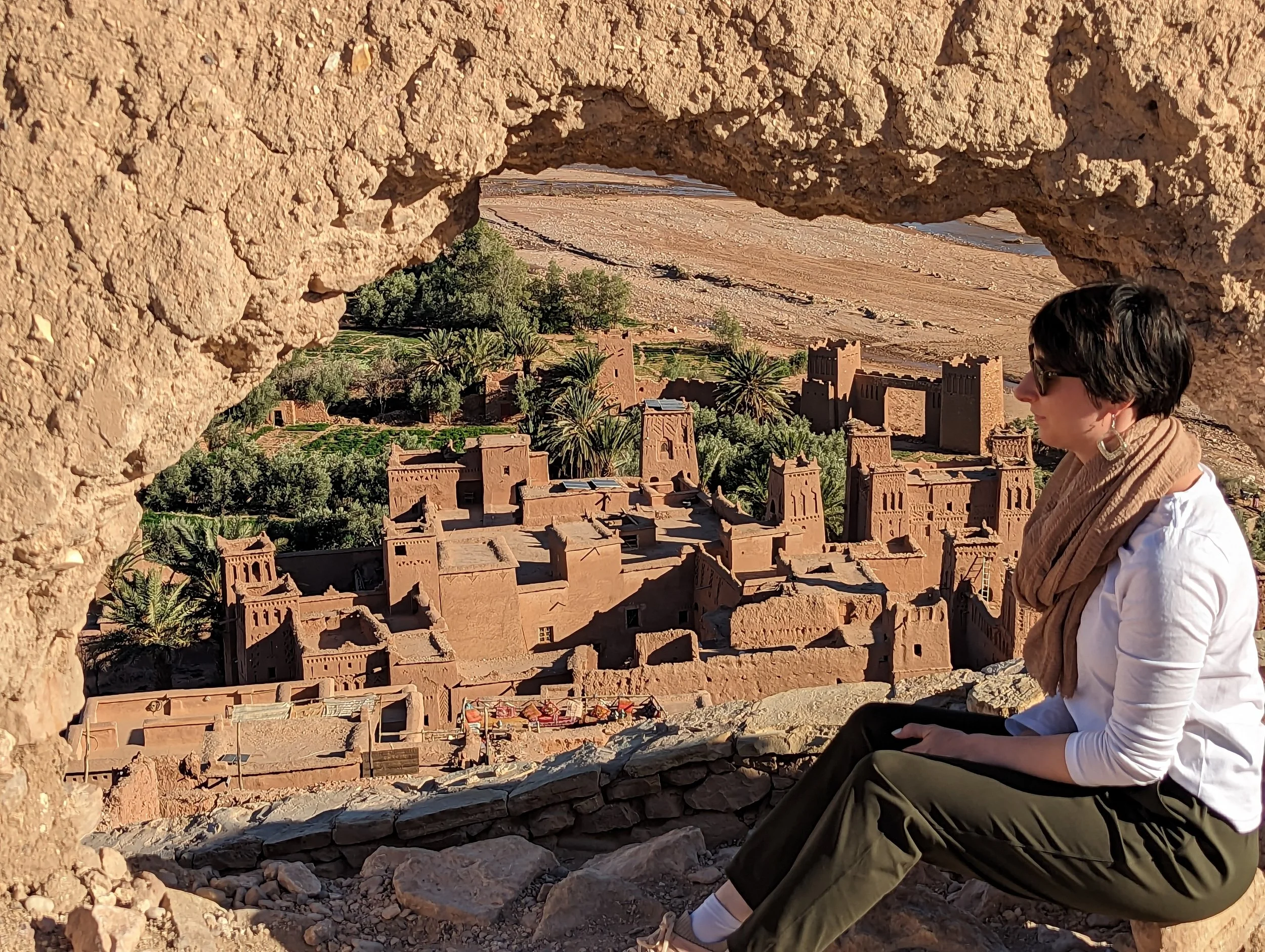 A woman sitting on a stone ledge, viewing a historic fortified village with terracotta buildings and palm trees through a rock arch.