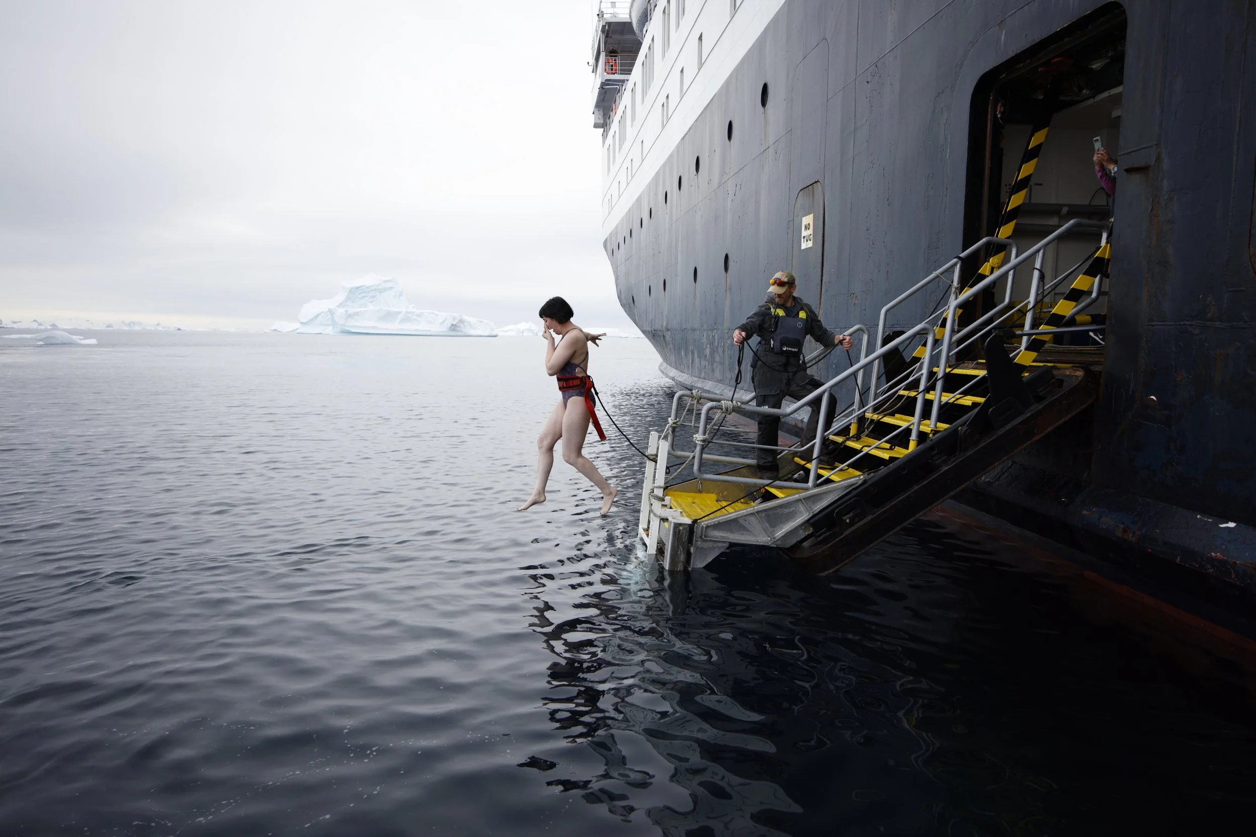 A woman in a swimsuit is jumping into icy water from a ship's platform, with a crew member assisting nearby, in a cold polar environment with icebergs in the background.