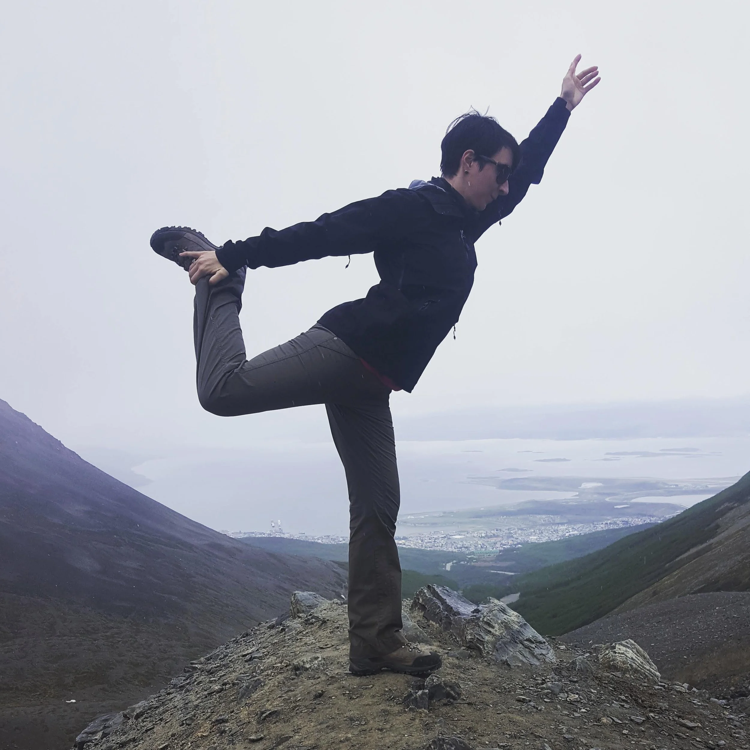 Person practicing yoga on rocky mountain trail overlooking a valley with water and town below, wearing black jacket, gray pants, and sunglasses, balancing in a yoga pose.