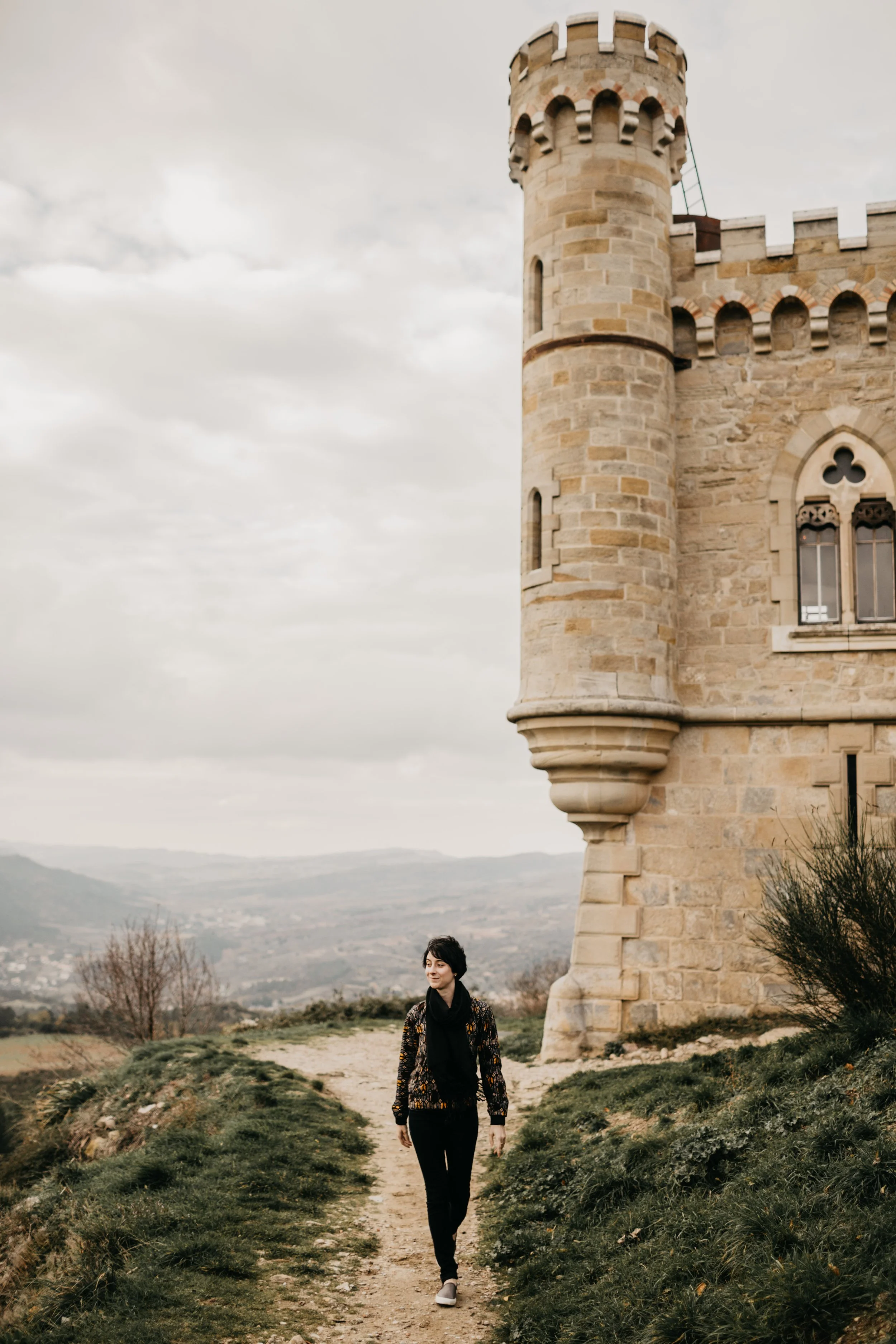 A woman walking on a dirt path near a castle tower with cloudy sky in the background.