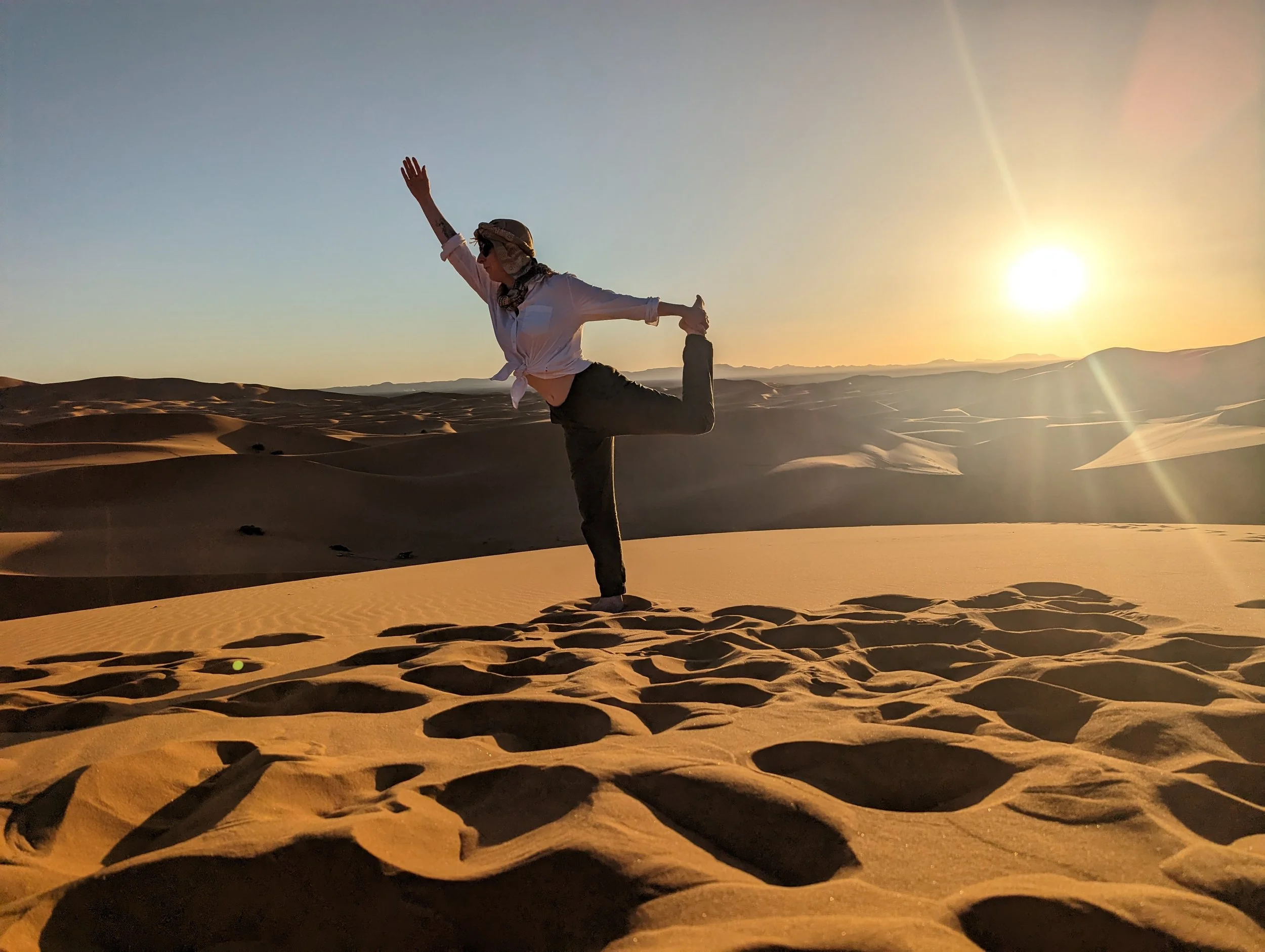 A person standing on sand dunes during sunset, performing a yoga pose with one leg bent and foot held behind, arm extended upward in a desert landscape.