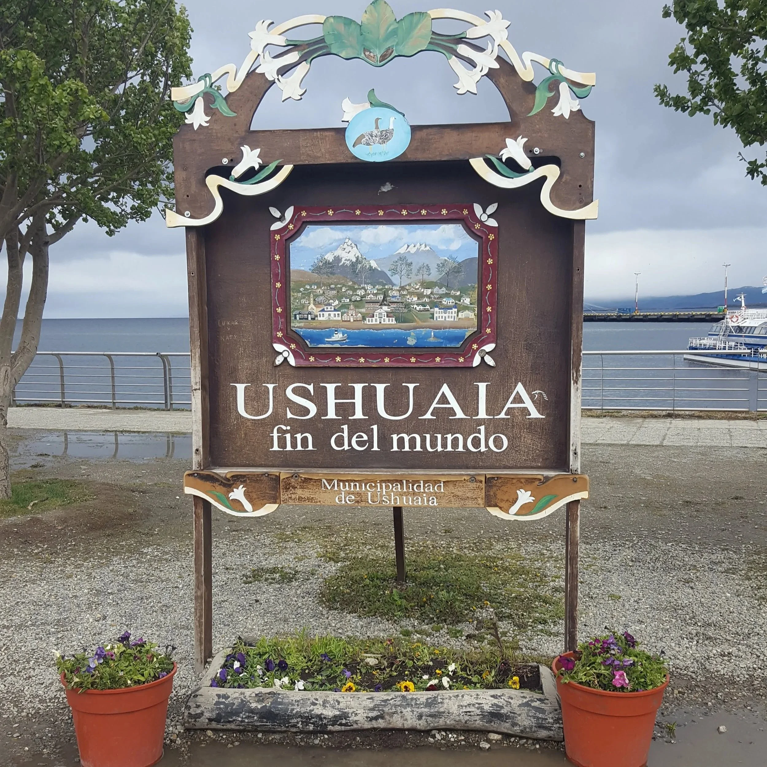 Sign for Ushuaia, Argentina, with mountain and cityscape illustration, and flower pots at the base, near the waterfront with boats and cloudy sky in the background.