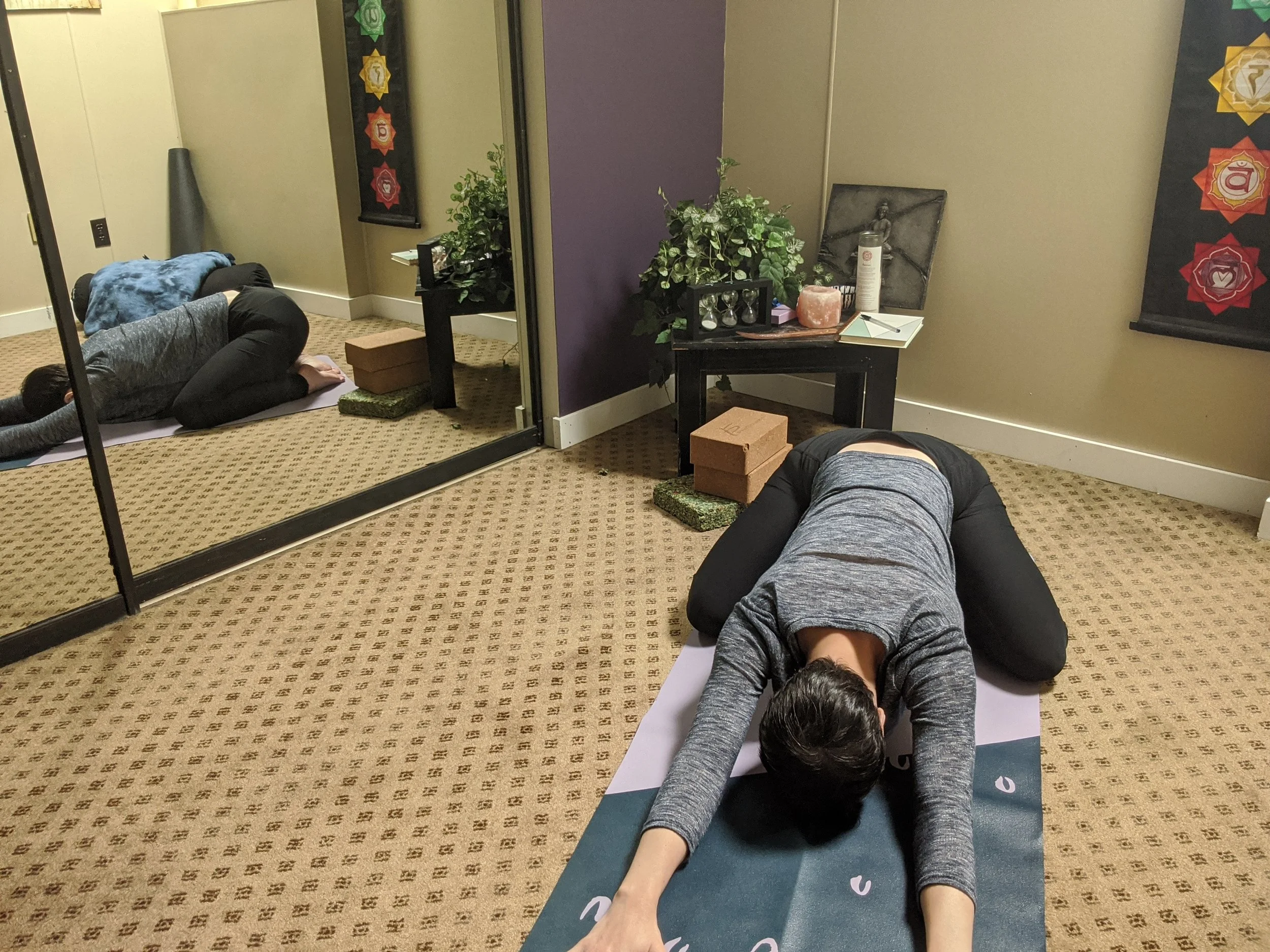 Person practicing yoga on a mat, in child's pose, in a room with a mirror, plants, and chakra-themed wall hangings.
