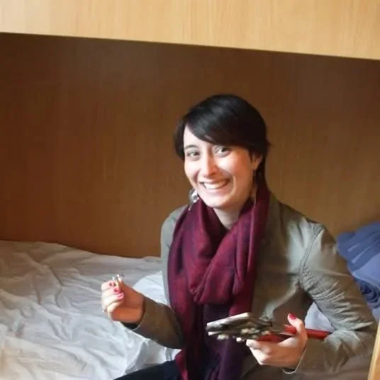 A woman with short dark hair, wearing a gray jacket and a maroon scarf, sitting on a bed in a room with a wooden headboard, smiling and holding a lock.