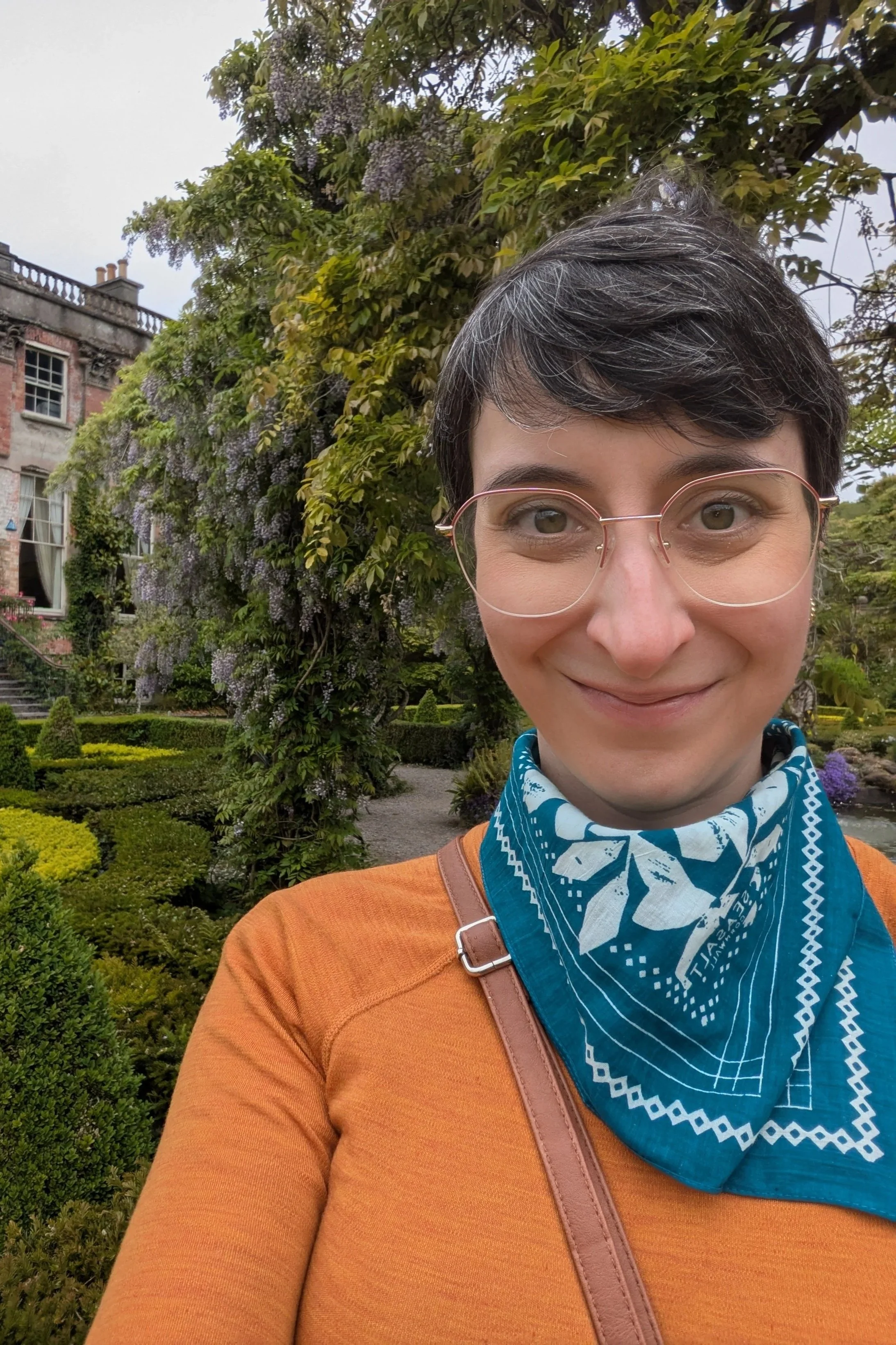 A woman smiling outdoors in a garden with lush greenery and flowering plants, wearing glasses, an orange top, and a blue bandana around her neck.