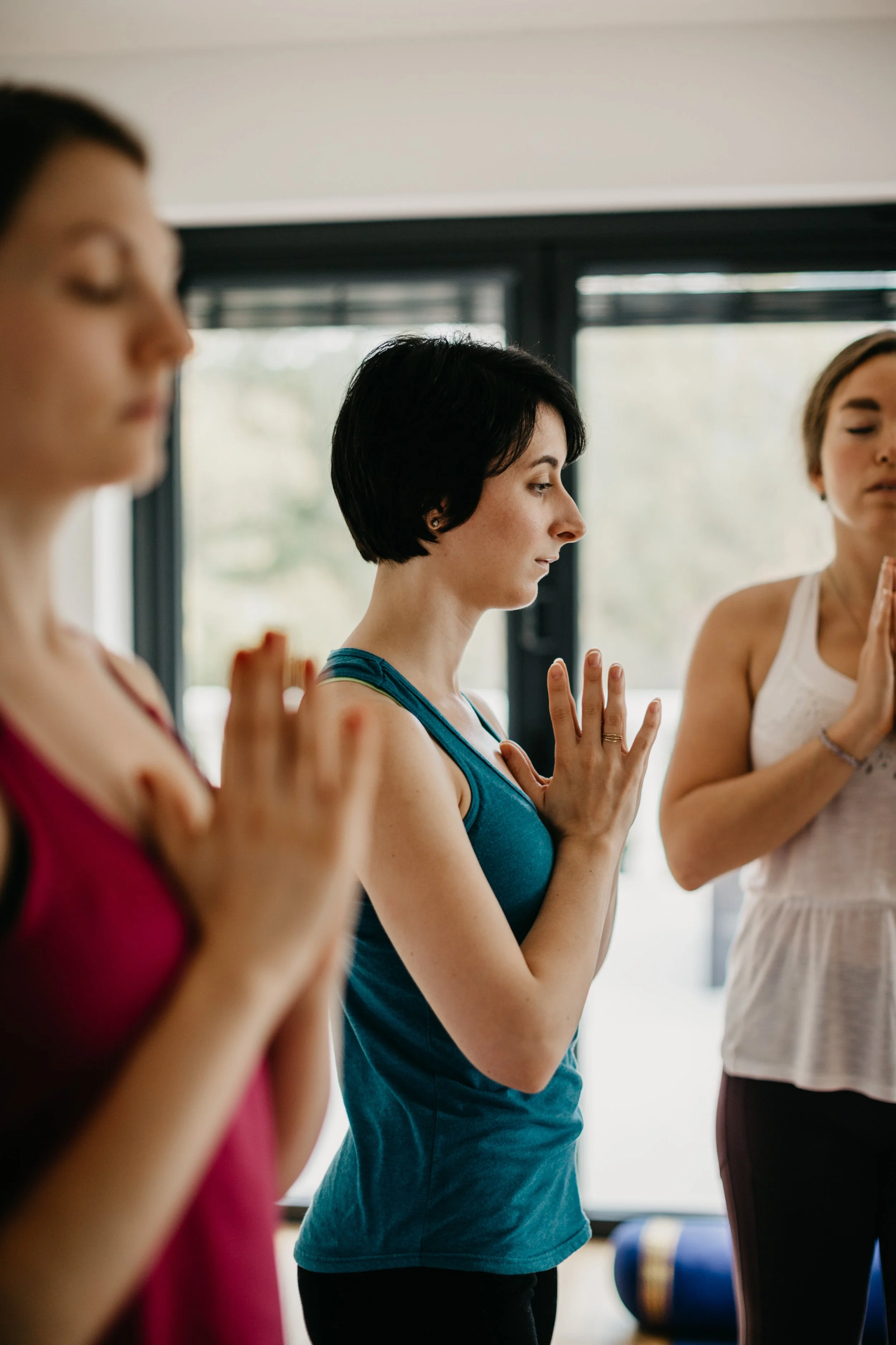 Three women practicing yoga indoors with eyes closed and hands in prayer position.