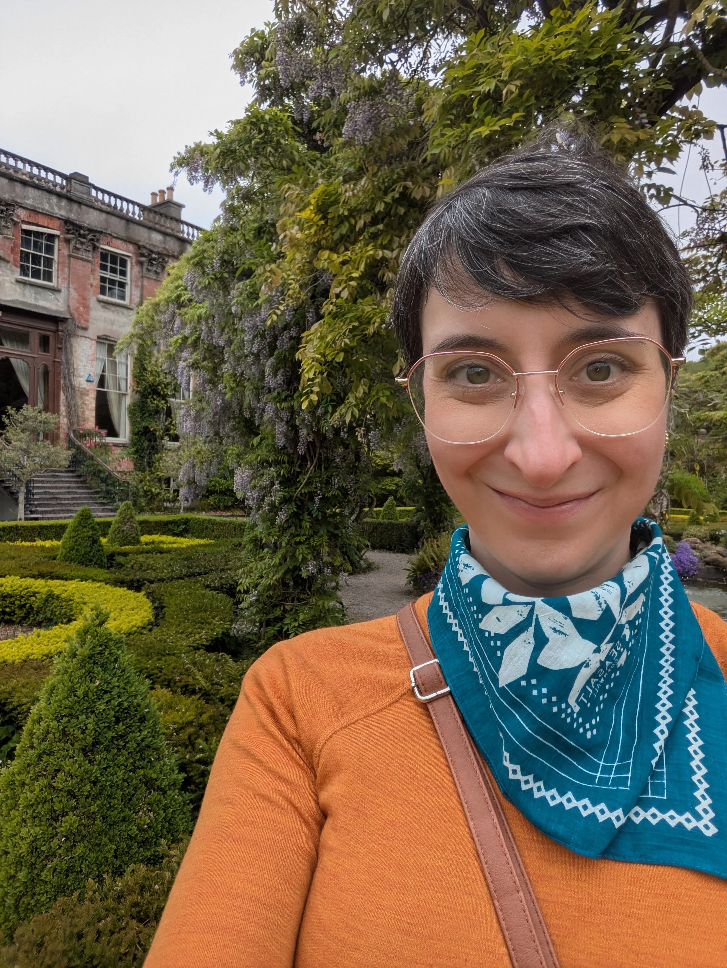 A woman with short dark hair and glasses smiling for a selfie outdoors amid a lush garden with green bushes, a flowering vine, and an old brick building in the background.