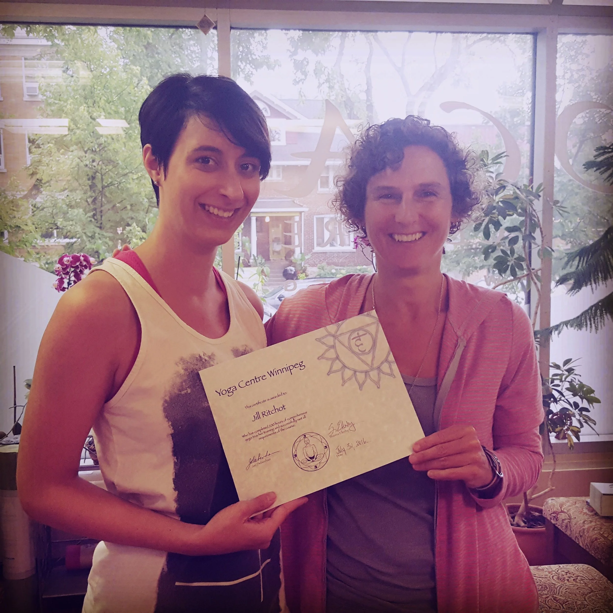 Two women standing indoors, smiling and holding a certificate from Yoga Centre Winnipeg. The woman on the left has short dark hair and wears a sleeveless top, while the woman on the right has curly hair and wears a pink zip-up jacket.