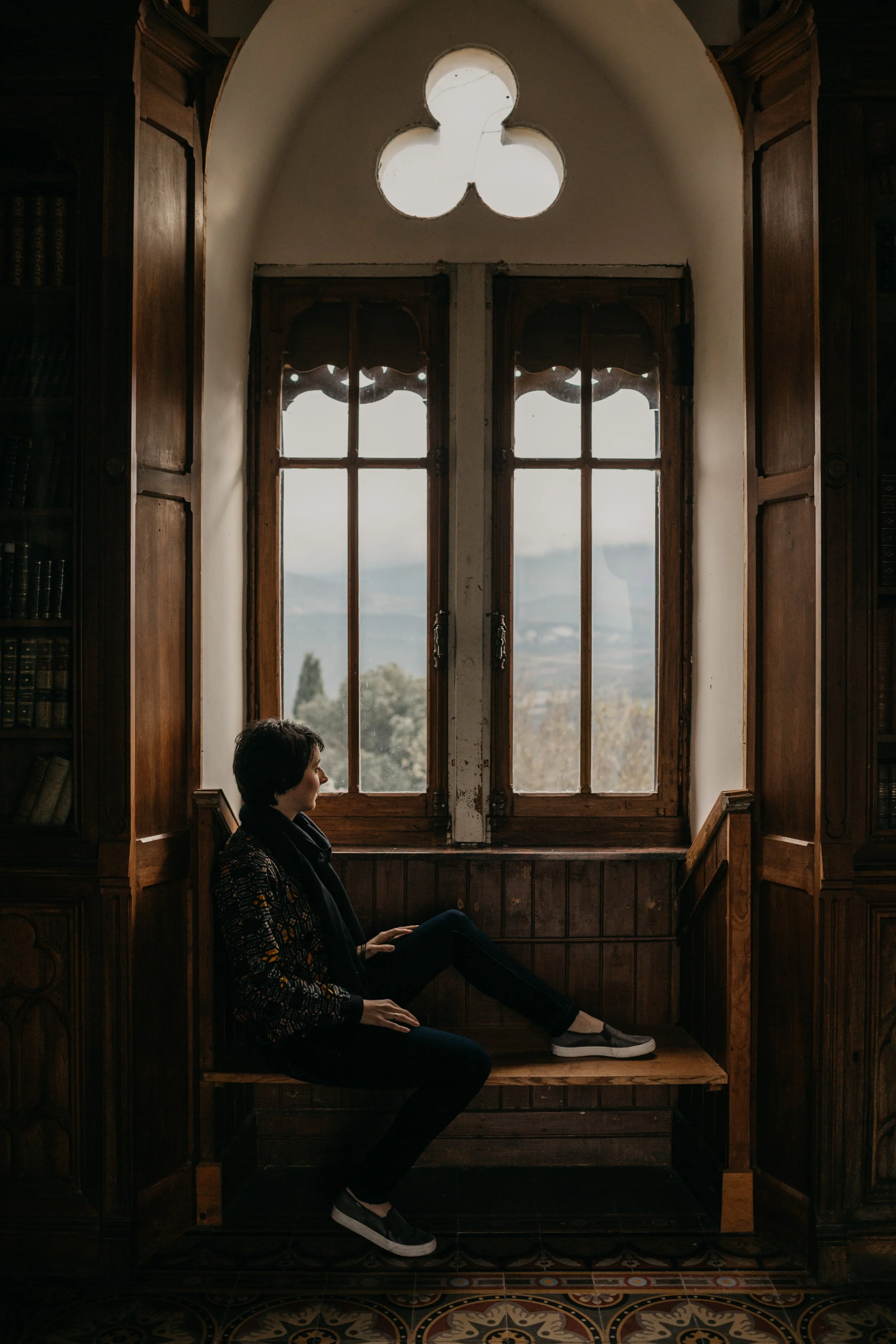 A woman sitting on a wooden bench in front of a large window with wooden frames inside a room with dark wood paneled walls and shelves. The window has a distinctive three-lobed shape at the top, and outside the window, a landscape with trees and mountains is visible.