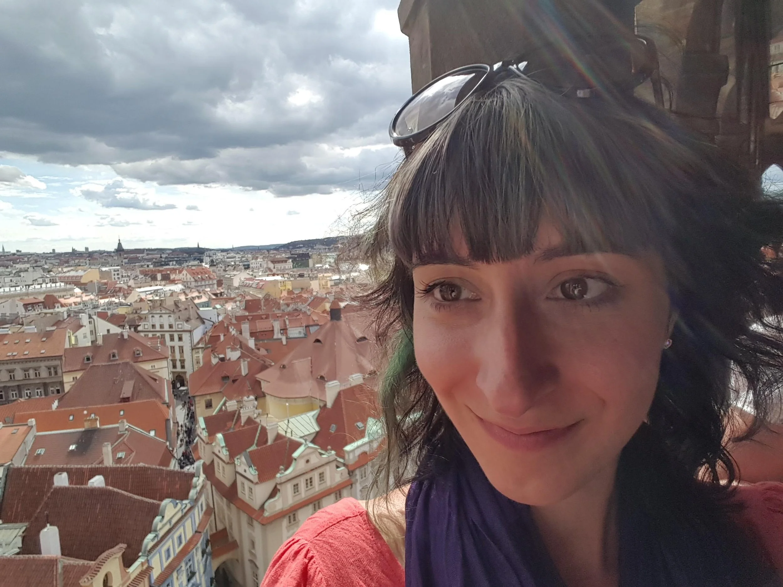 A woman with short dark hair and sunglasses on her head, smiling, with a cityscape of red-tiled rooftops and historic buildings under a partly cloudy sky in the background.