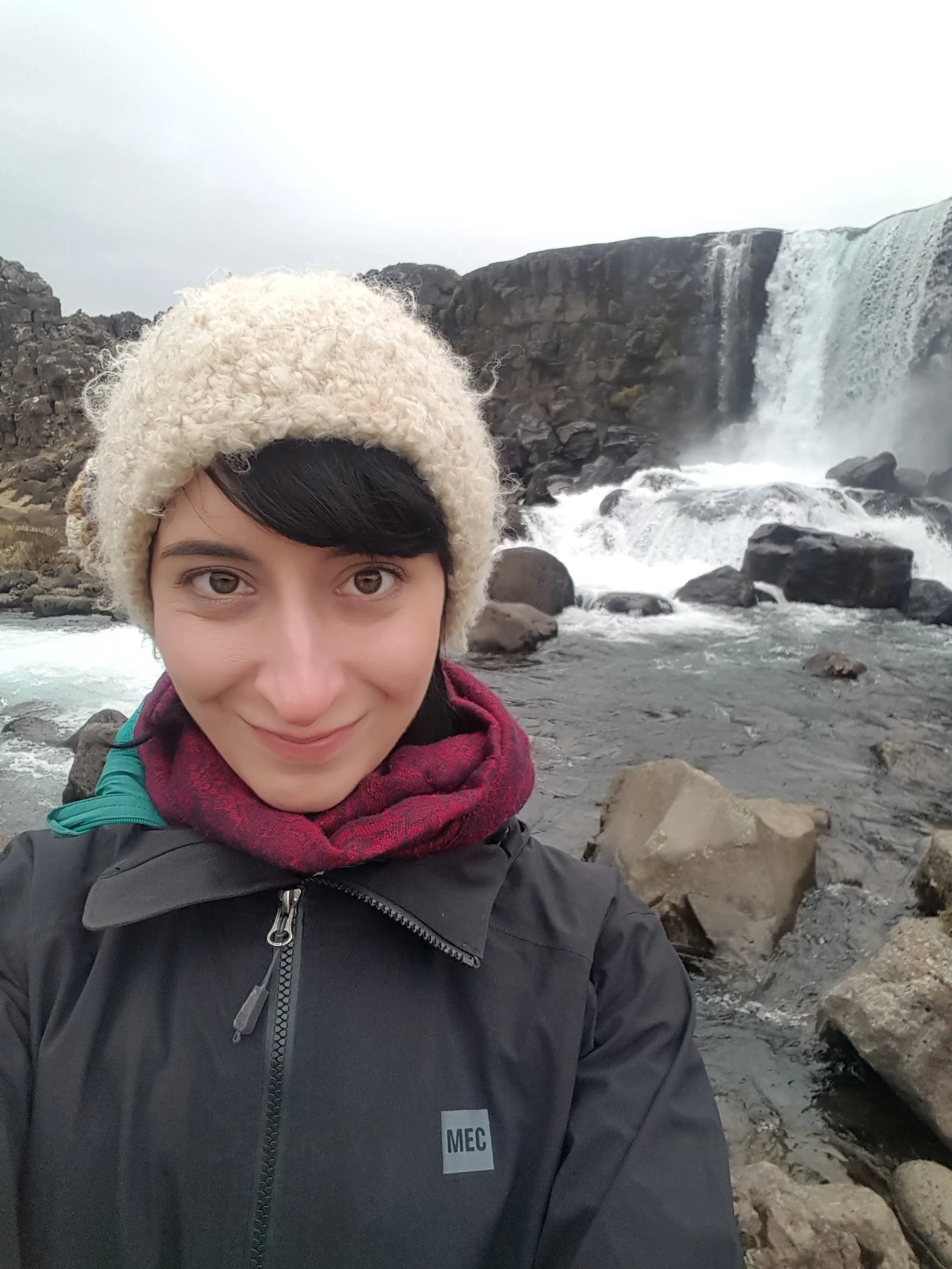 A woman taking a selfie in front of a waterfall surrounded by rocks, wearing a beige knit hat, a dark jacket, and a red scarf.