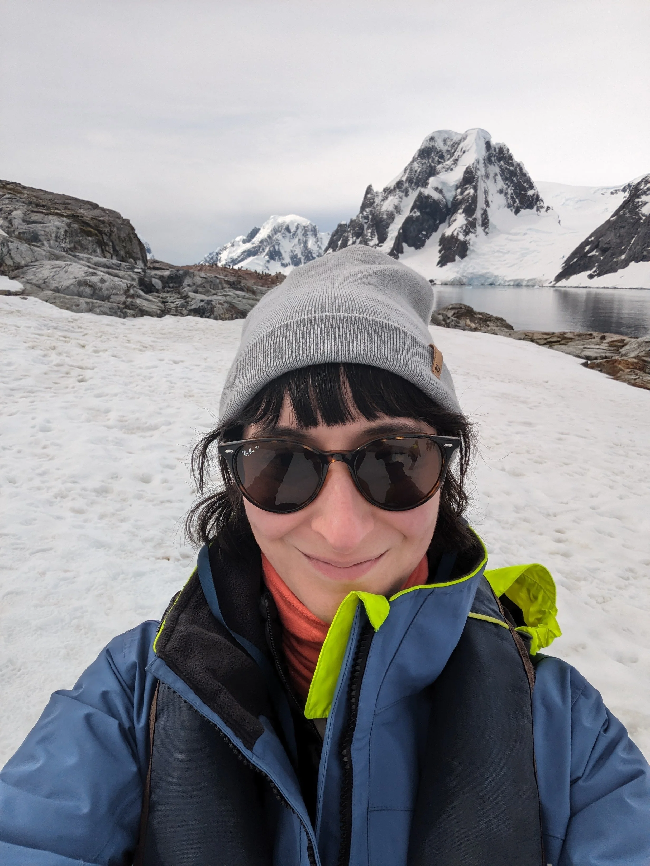 A person in outdoor winter gear taking a selfie in a snowy mountainous landscape with snow-covered peaks and a body of water in the background.