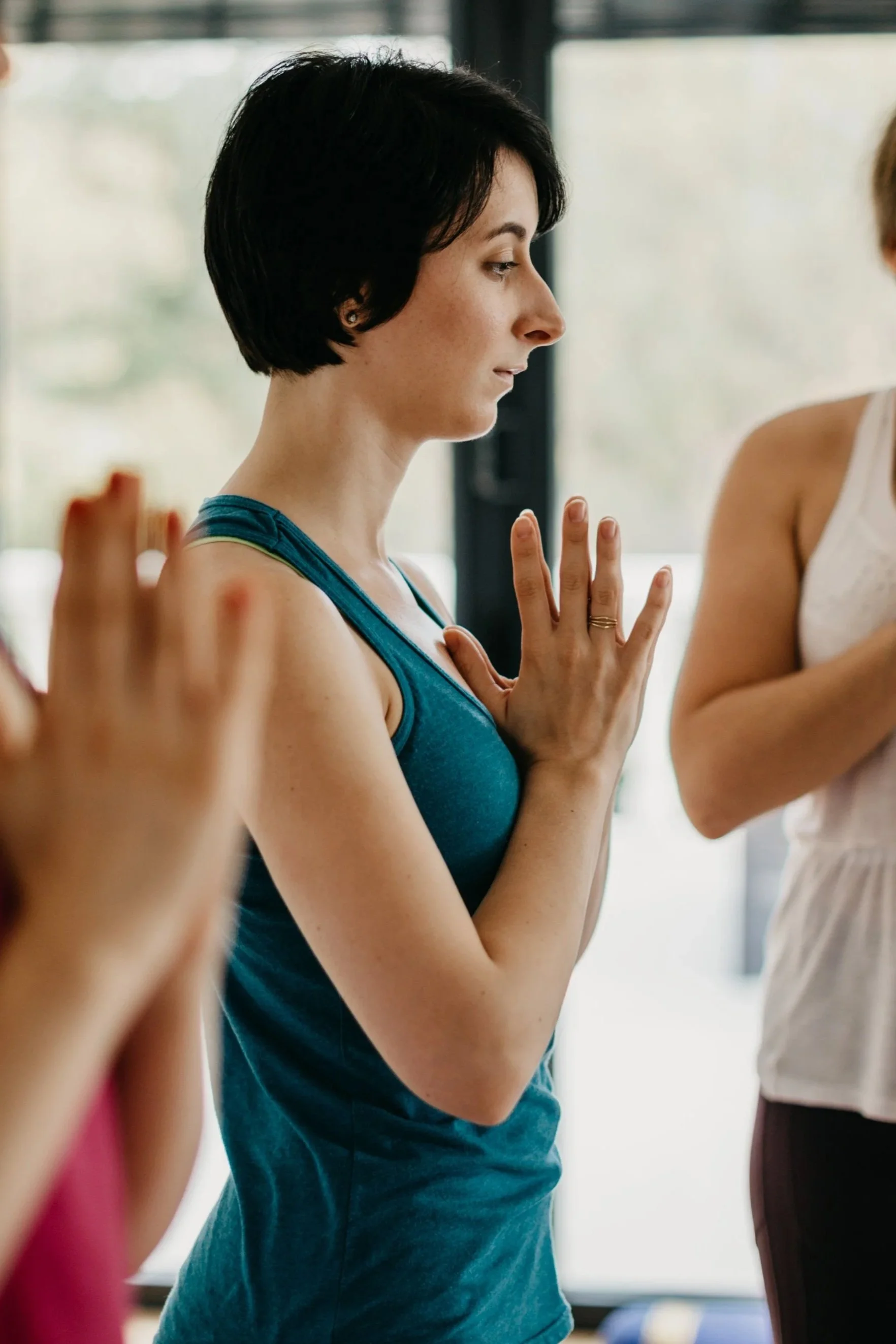 A woman with short dark hair in a teal tank top practicing yoga with eyes closed, hands pressed together at her chest, in a bright indoor space.