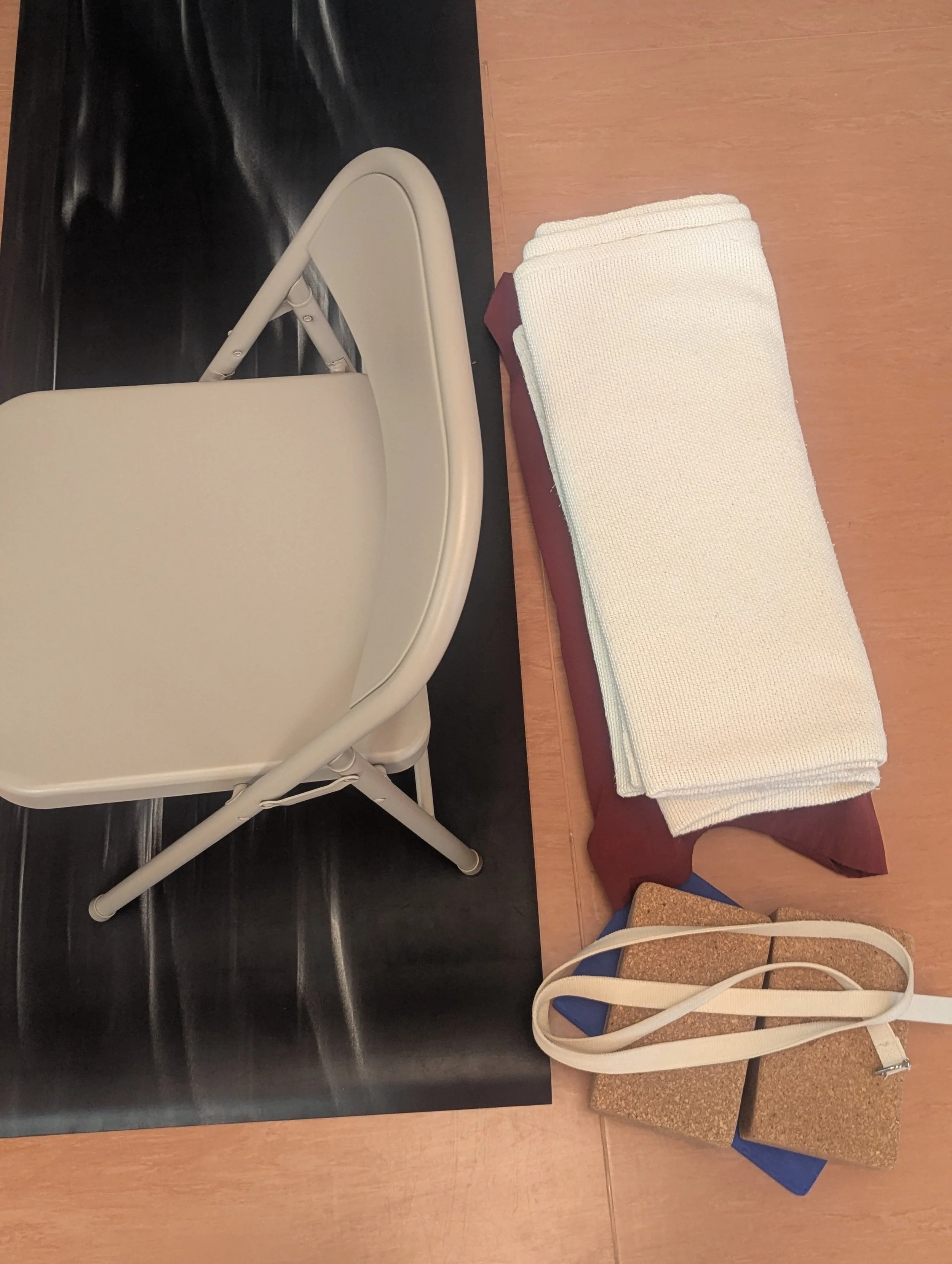 A beige folding chair, a folded white cloth, a maroon cloth, cork yoga blocks, a beige yoga strap, and a black yoga block on a wooden floor.
